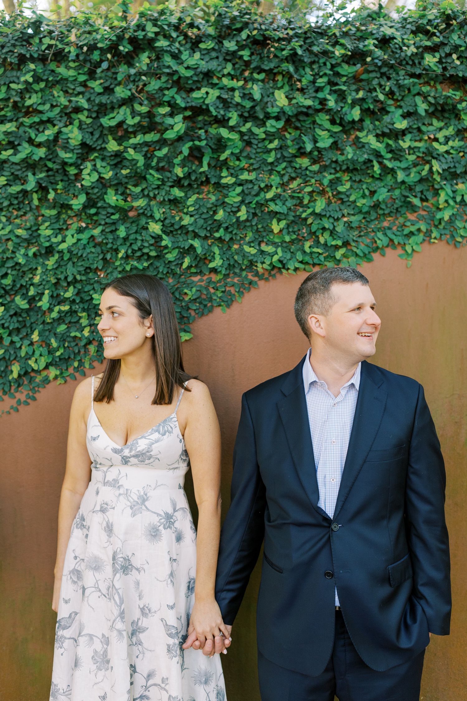A couple holds hands while standing against an ivy-covered wall, with the woman in a floral dress and man in navy suit.
