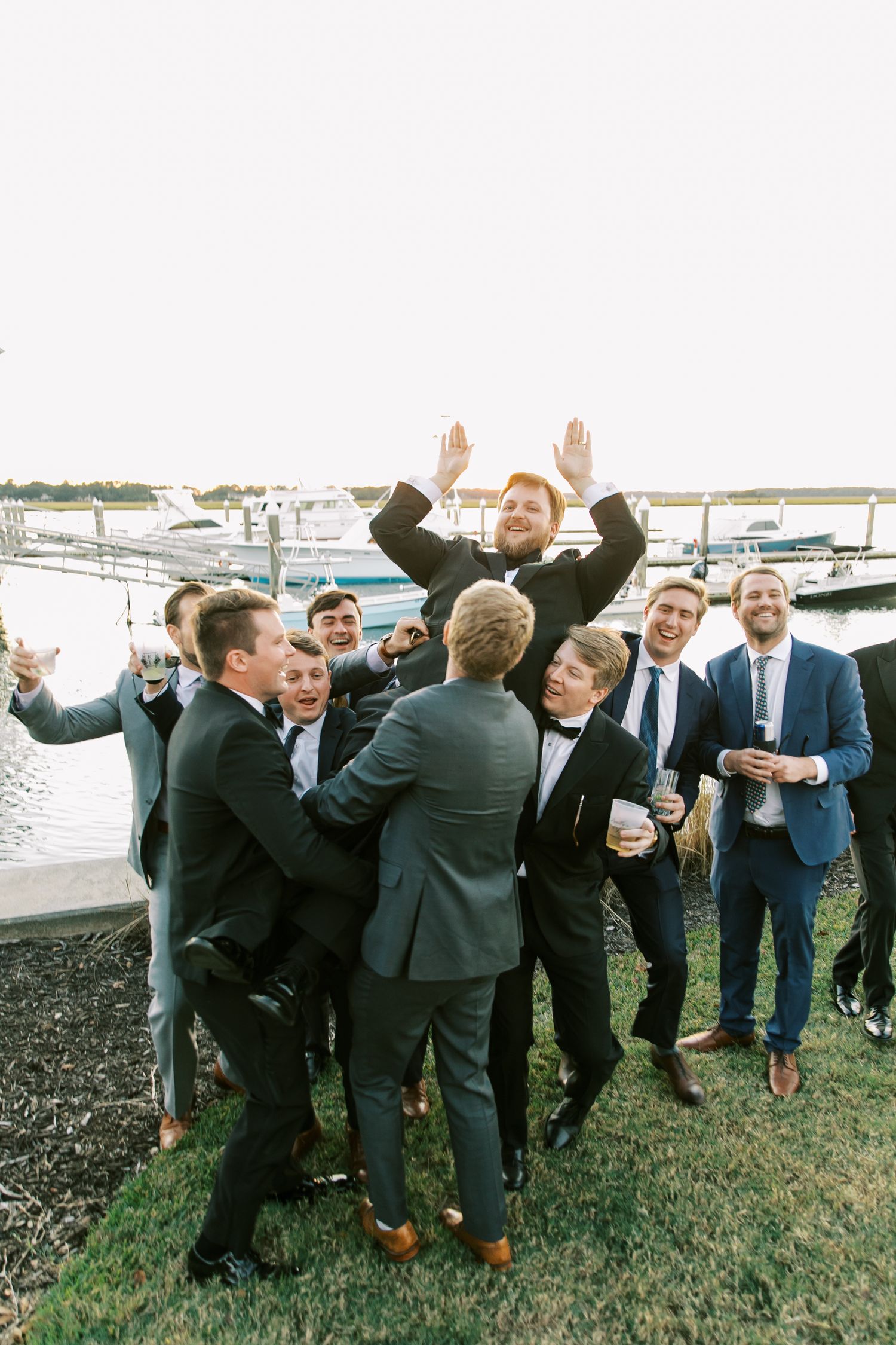 Groomsmen celebrating joyfully on waterfront lawn.