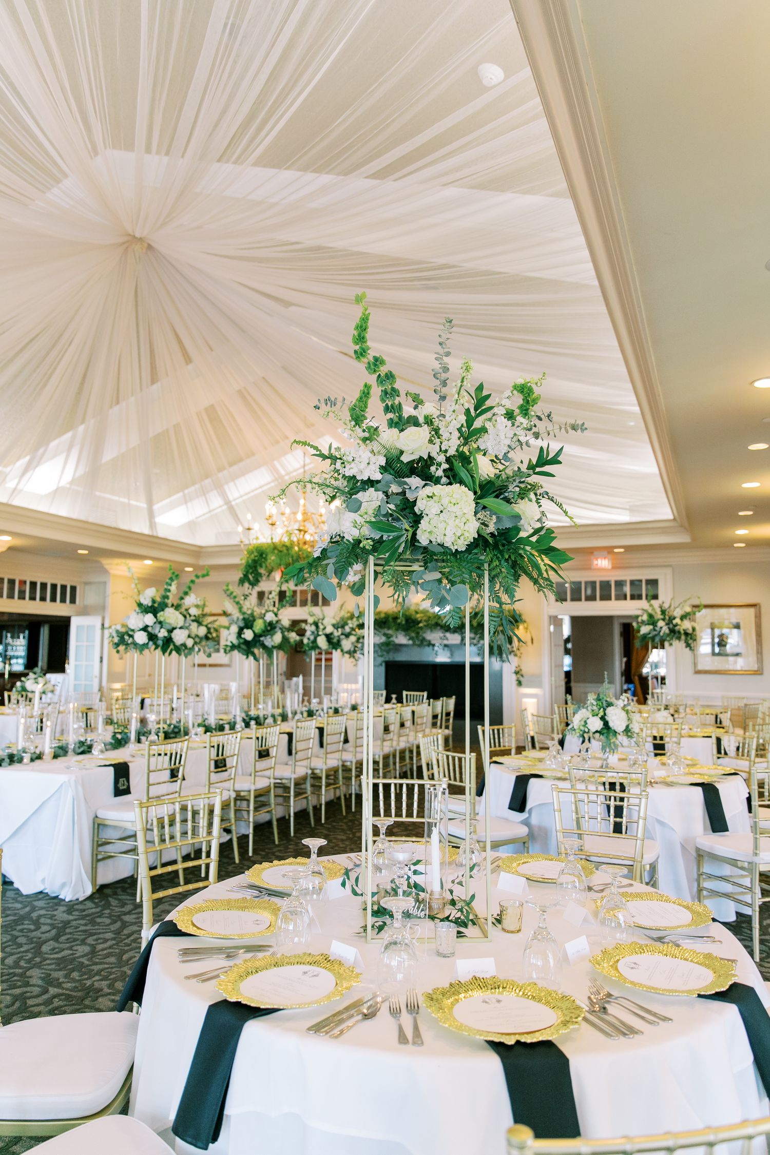 Elegant wedding reception venue with white draped ceiling and tall floral centerpieces featuring white flowers and greenery.
