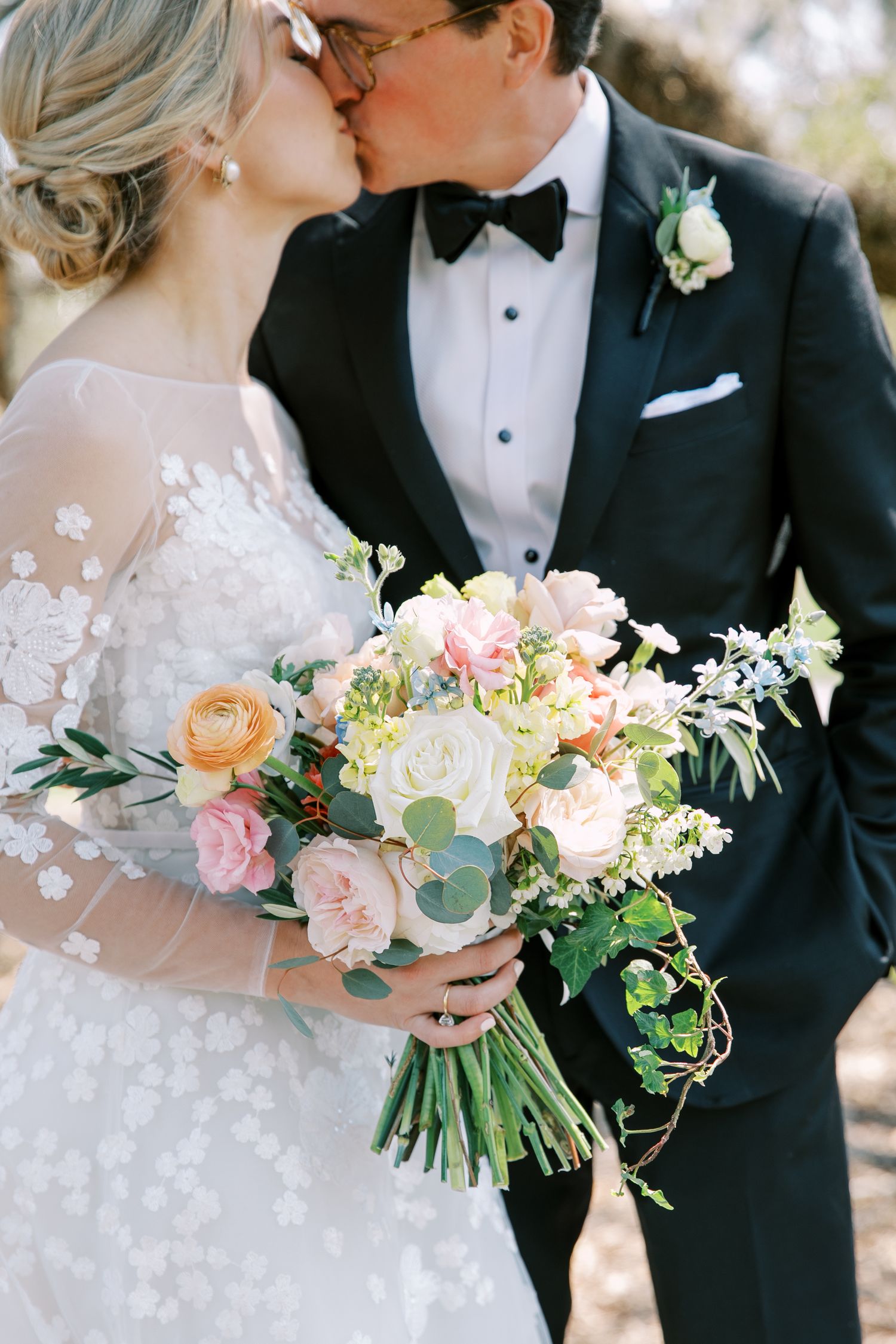 A romantic wedding couple shares a kiss while holding a lush bouquet of peach and pink roses with greenery.