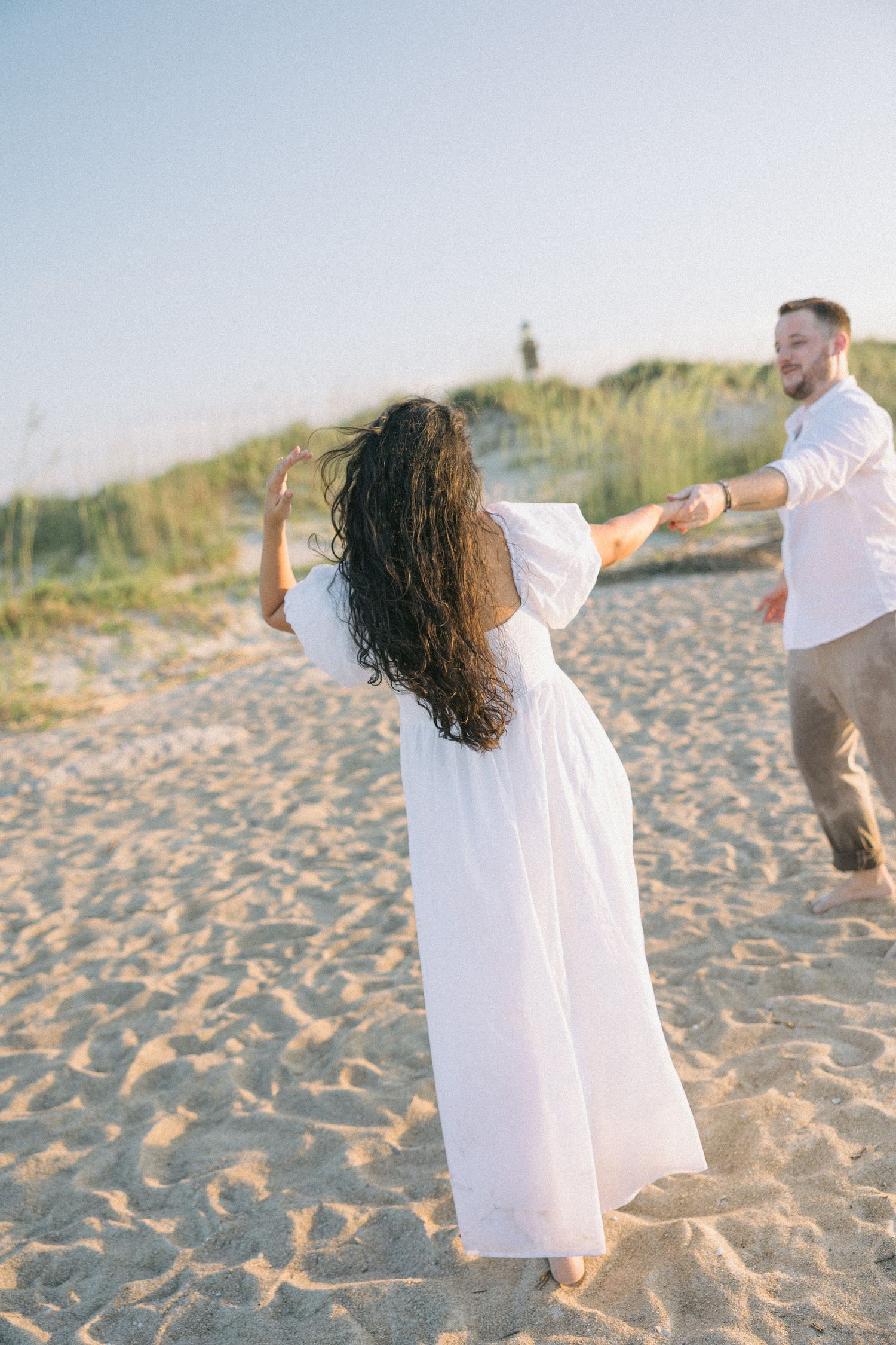 Two people in white clothing dance playfully on a sandy beach with sea grass in the background.