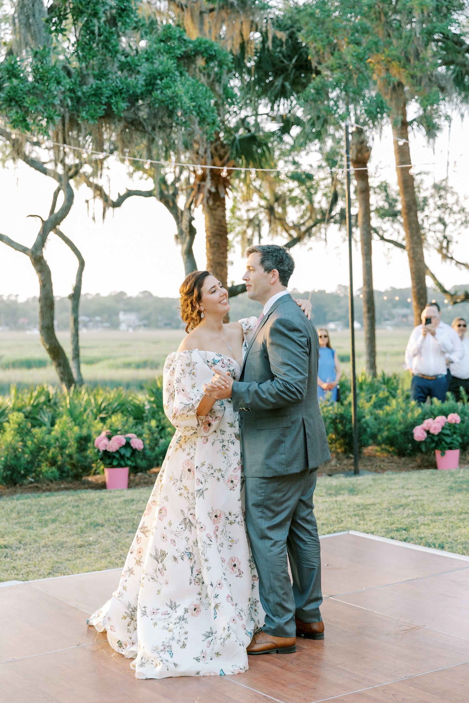 A couple dances outdoors on a patio with hanging lights and pink flowers in the background.