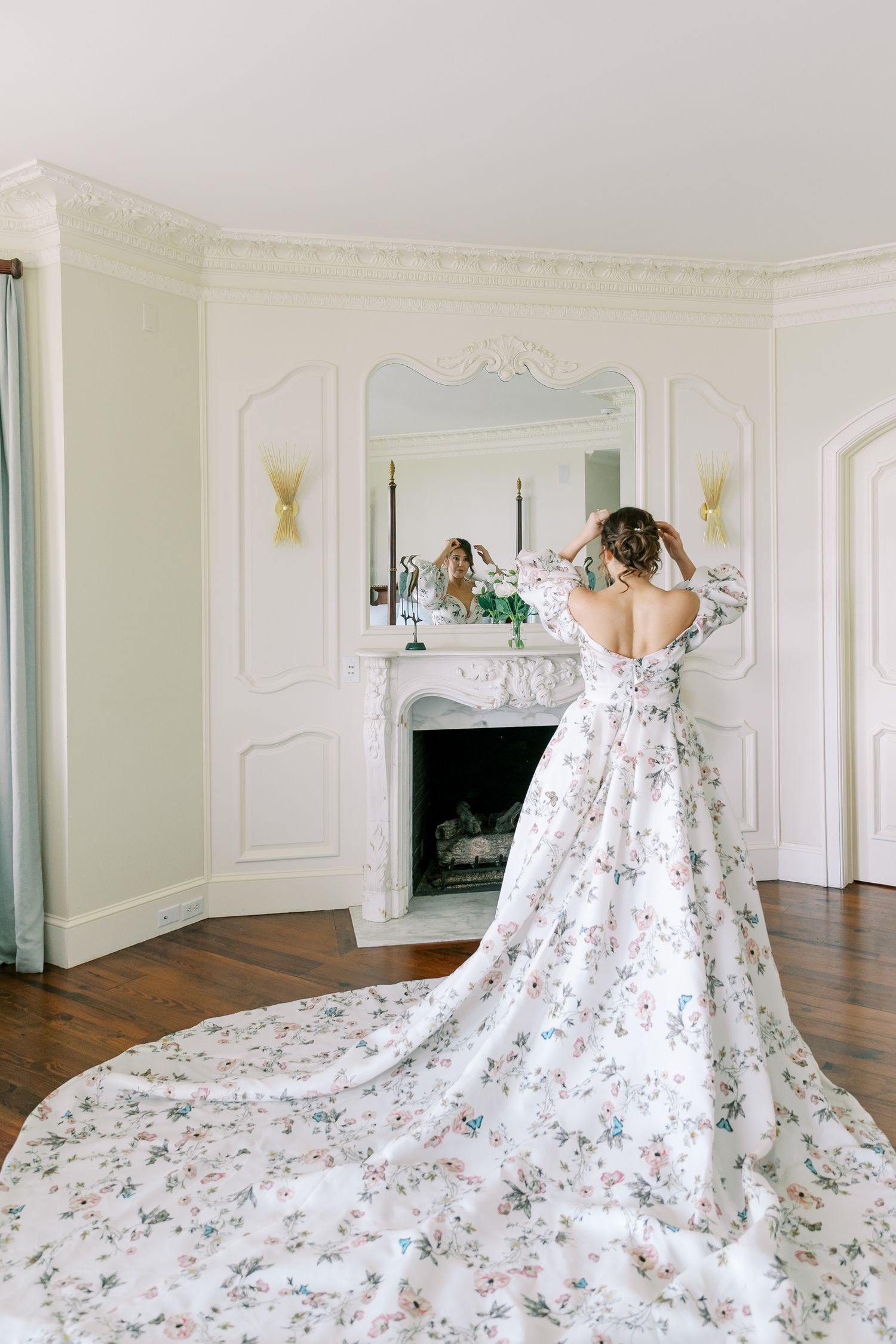 An elegant floral wedding gown with long train displayed in front of an ornate fireplace.