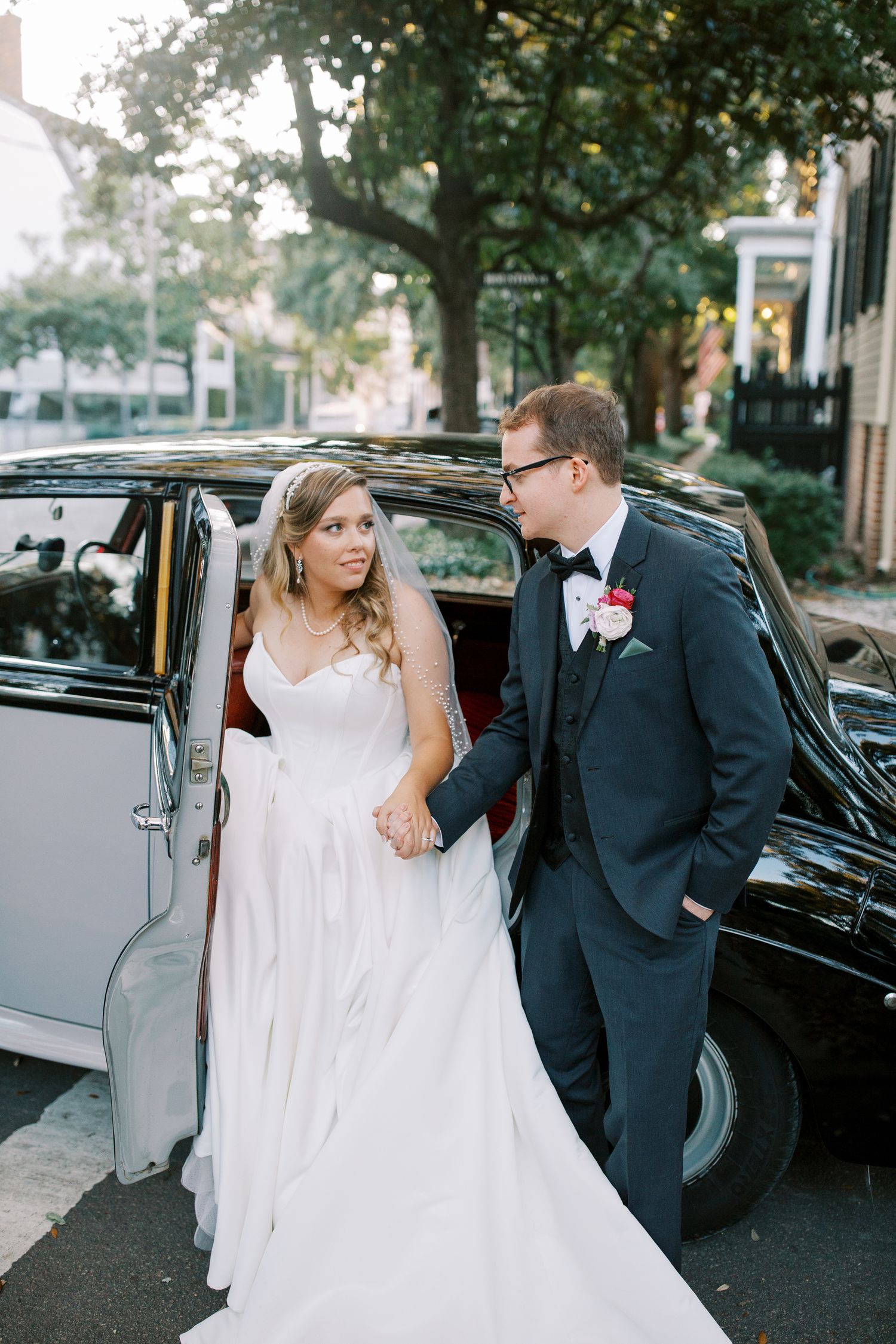 A bride and groom stand beside a classic vintage car on their wedding day, with trees in the background.