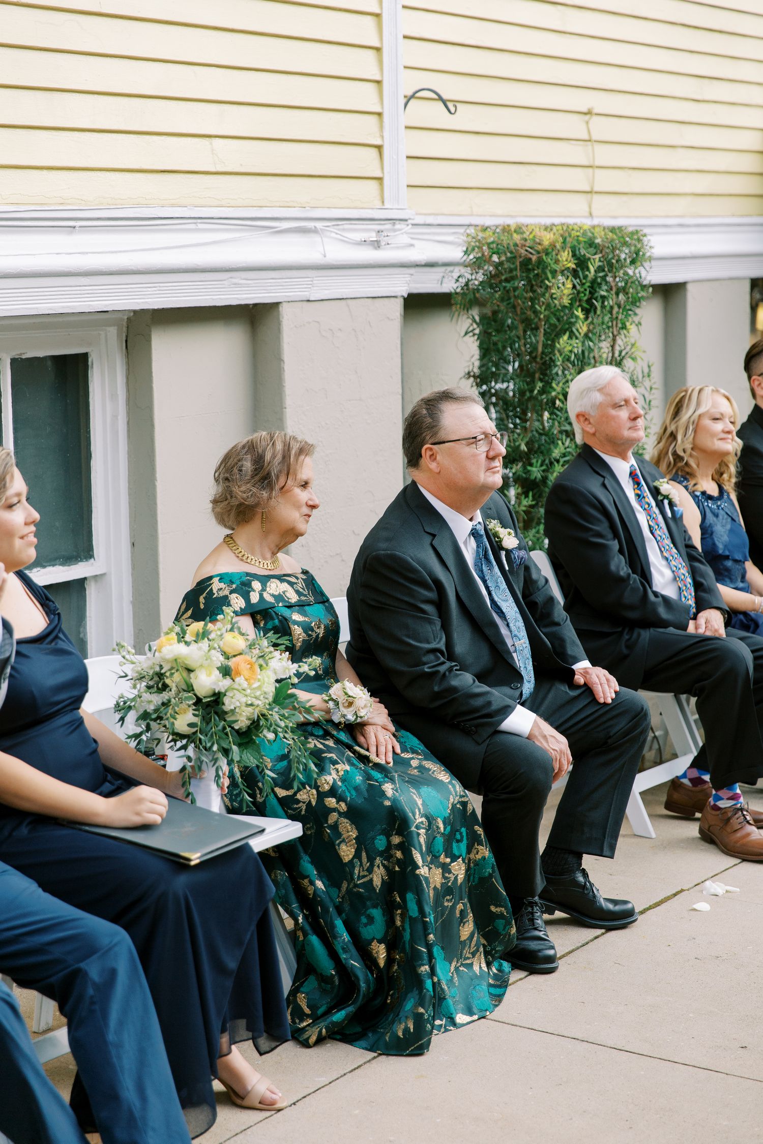 Wedding attendees in formal attire seated along a white building exterior.