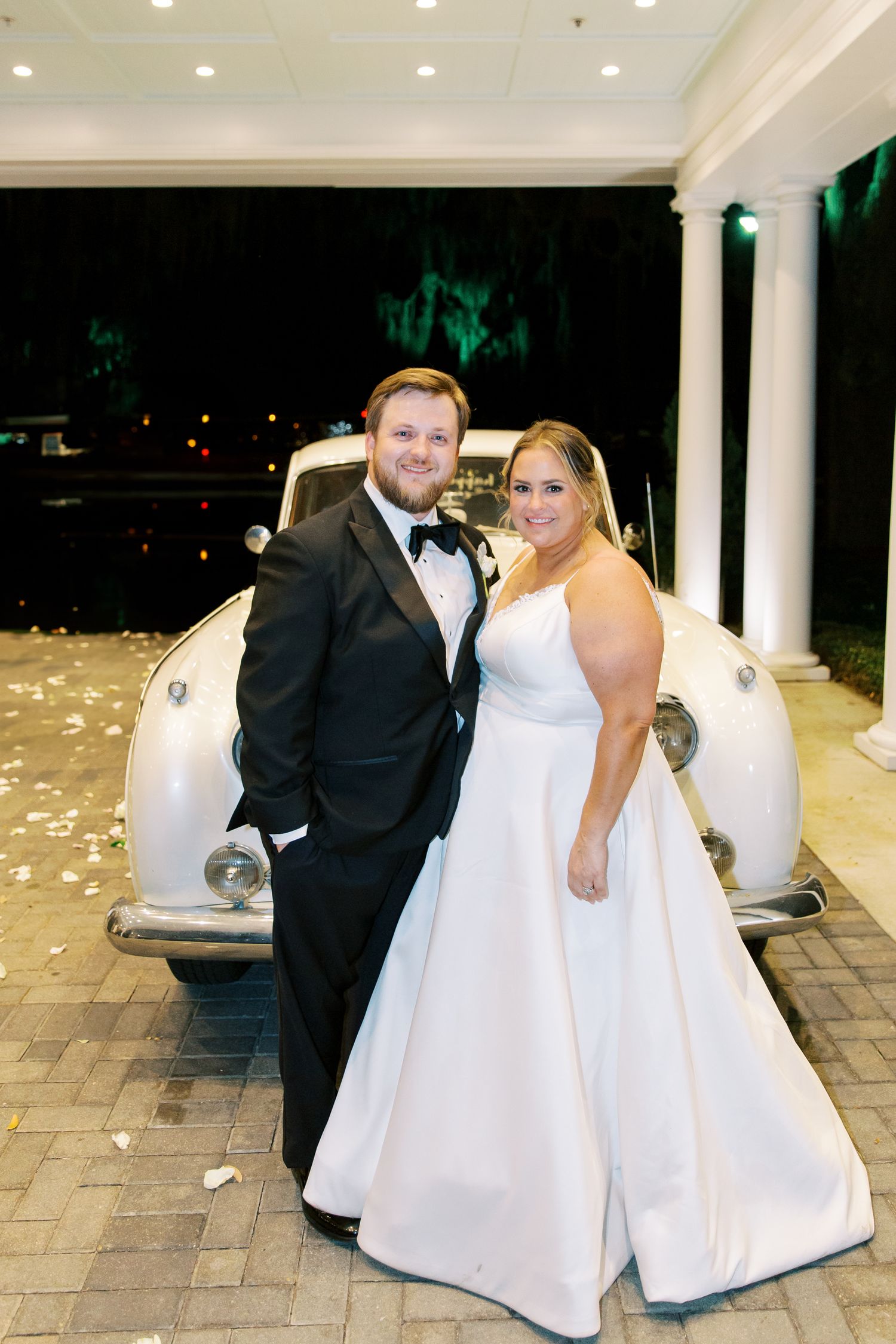 Elegant couple poses in formal attire next to vintage white car on their wedding night.