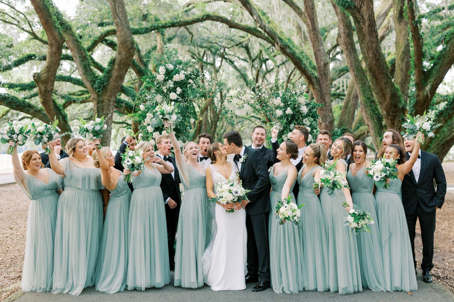 Wedding party in sage green dresses and black tuxedos posing under oak trees at Belfair in Bluffton.