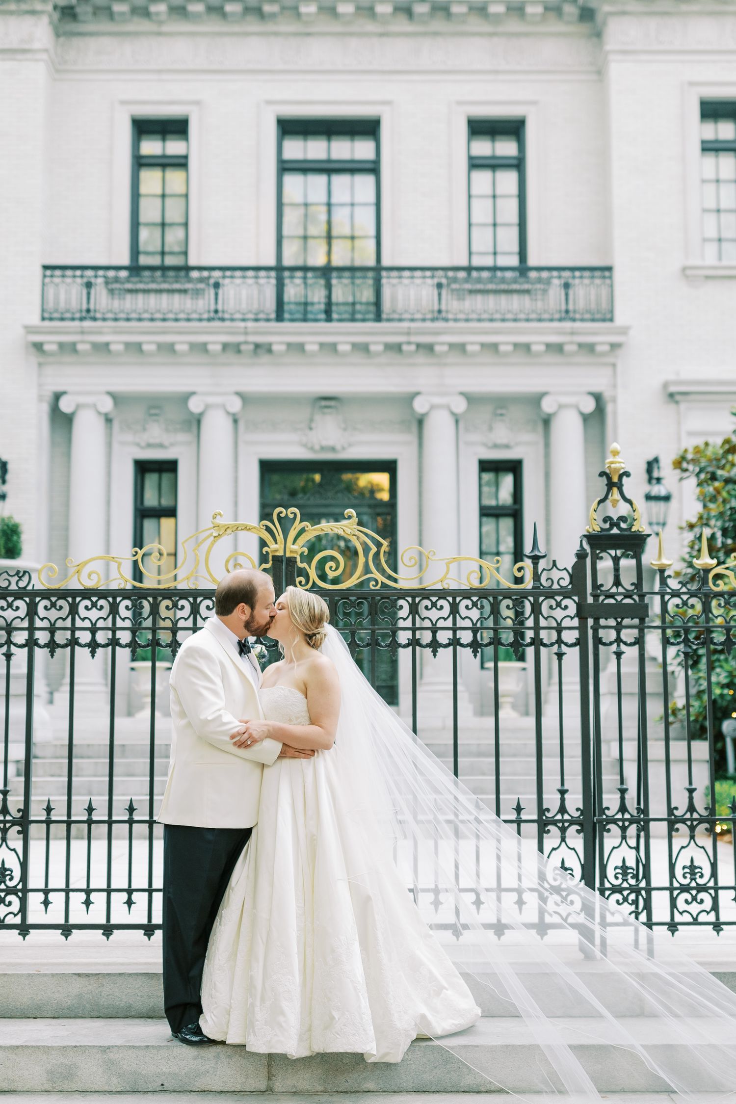 Elegant wedding portrait in front of historic white mansion with ornate iron gates.