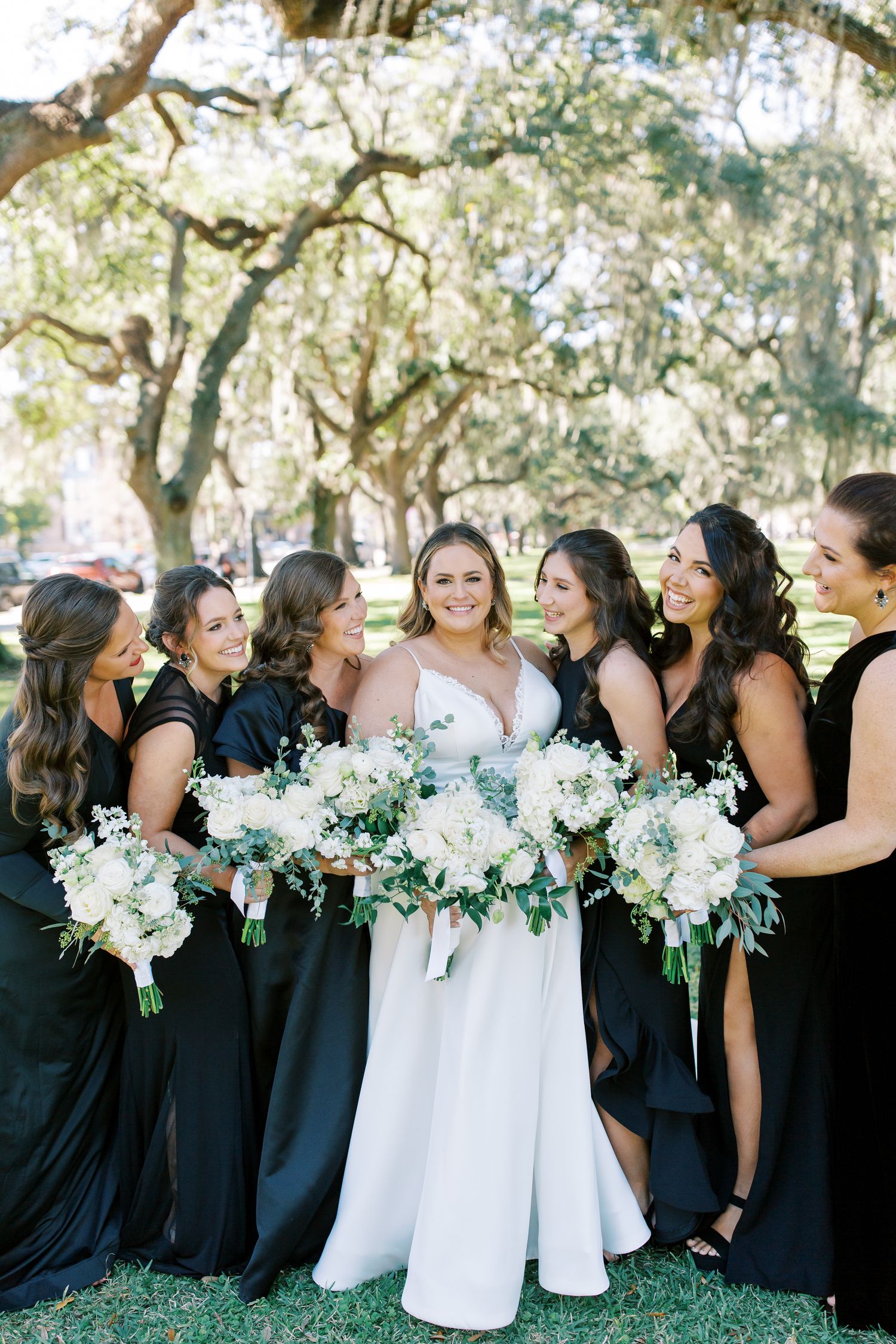 Bridesmaids in black dresses gather with bouquets under moss-draped trees.