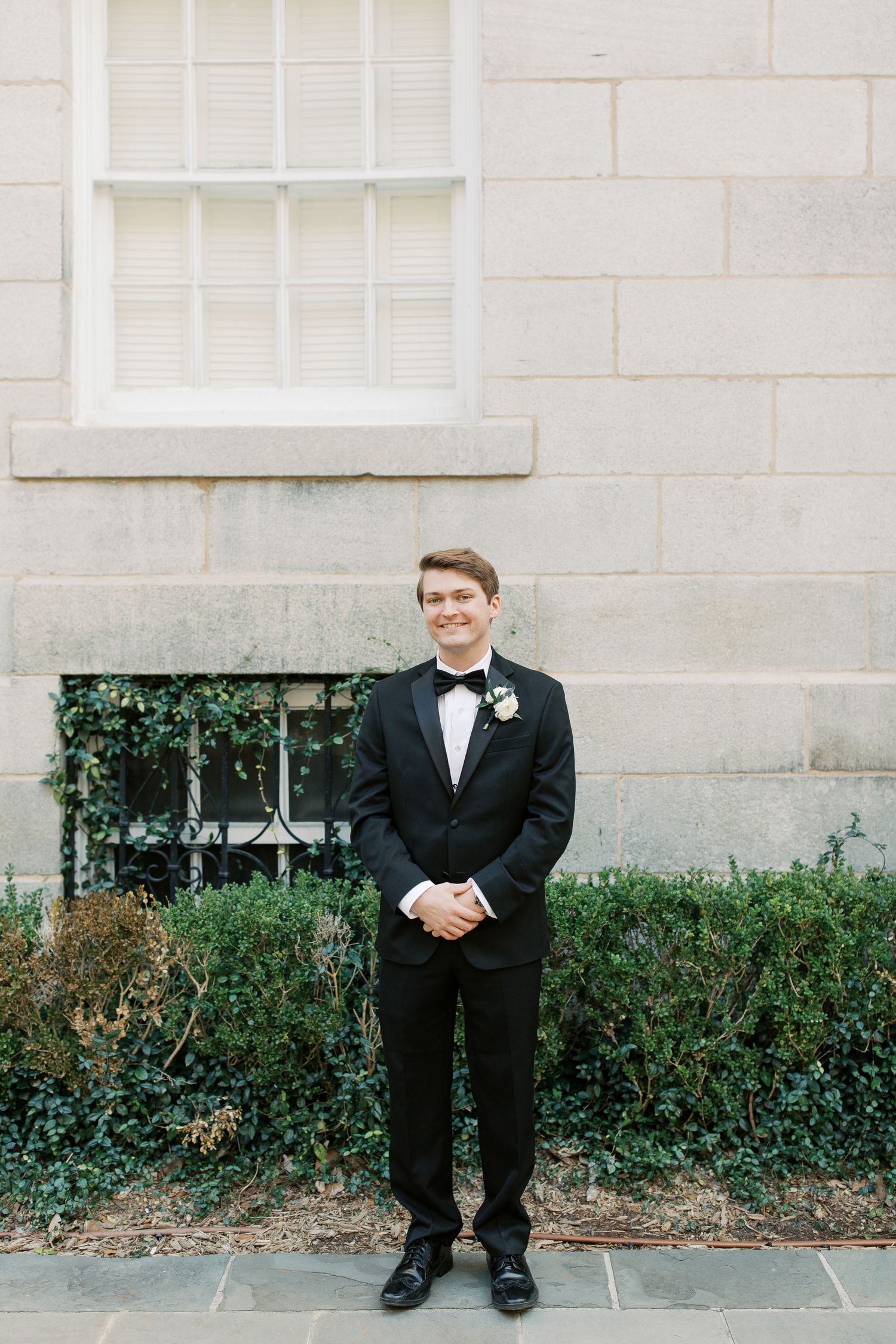 A groom in a classic black tuxedo and bowtie stands against a white stone wall with greenery.