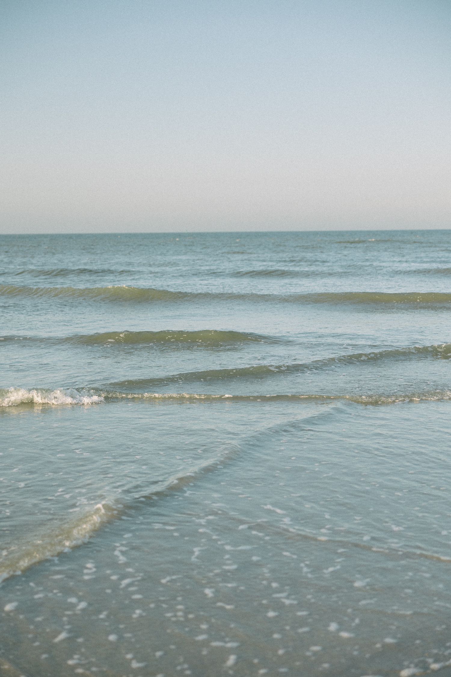 Gentle waves roll onto a sandy beach under a soft blue sky.
