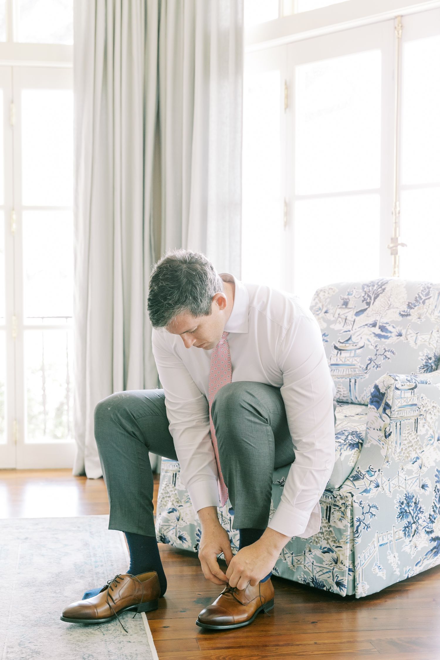 Person in white shirt and green pants tying brown leather dress shoes while sitting on a patterned armchair.