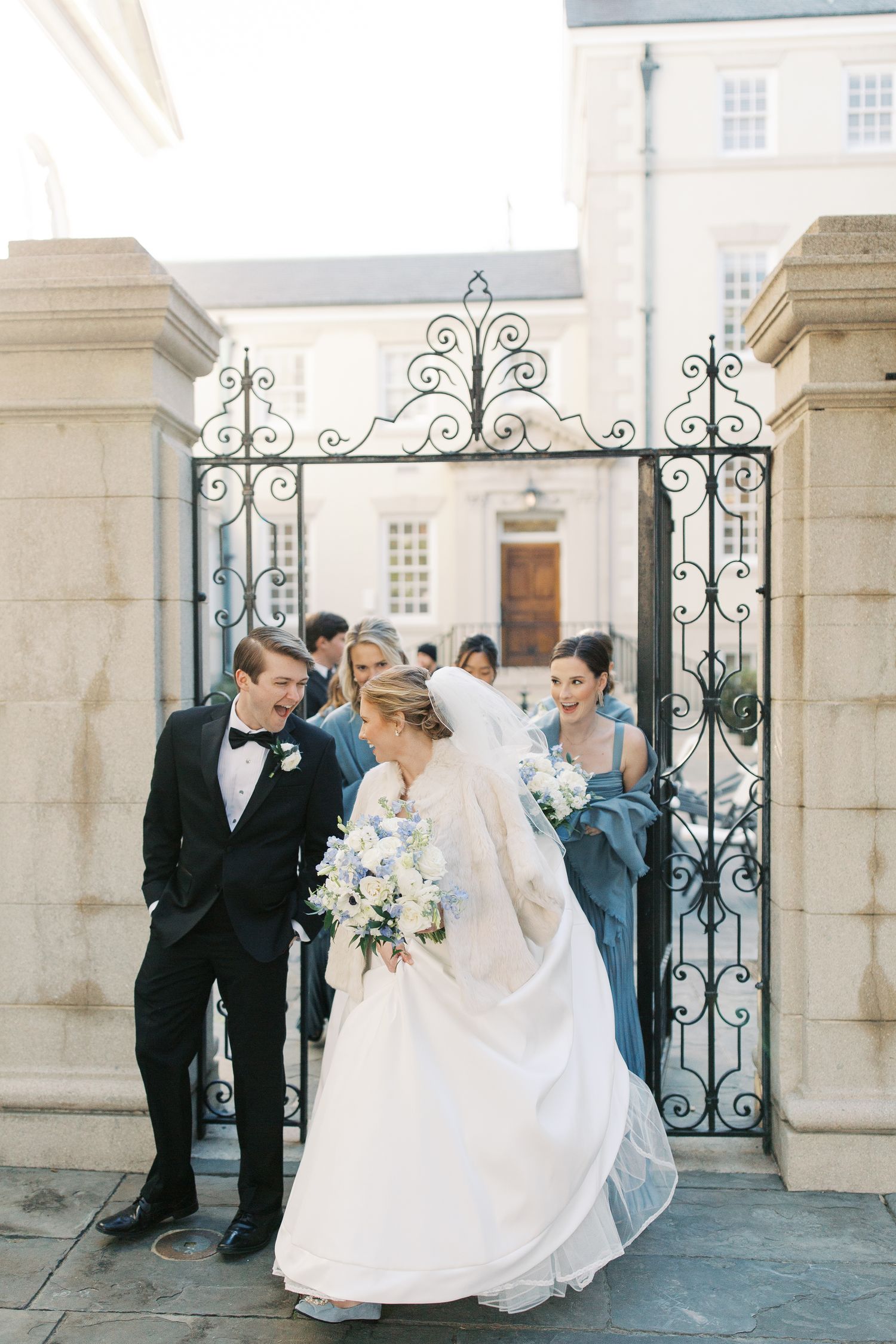 Wedding party gathered by ornate iron gates at an elegant stone building entrance.
