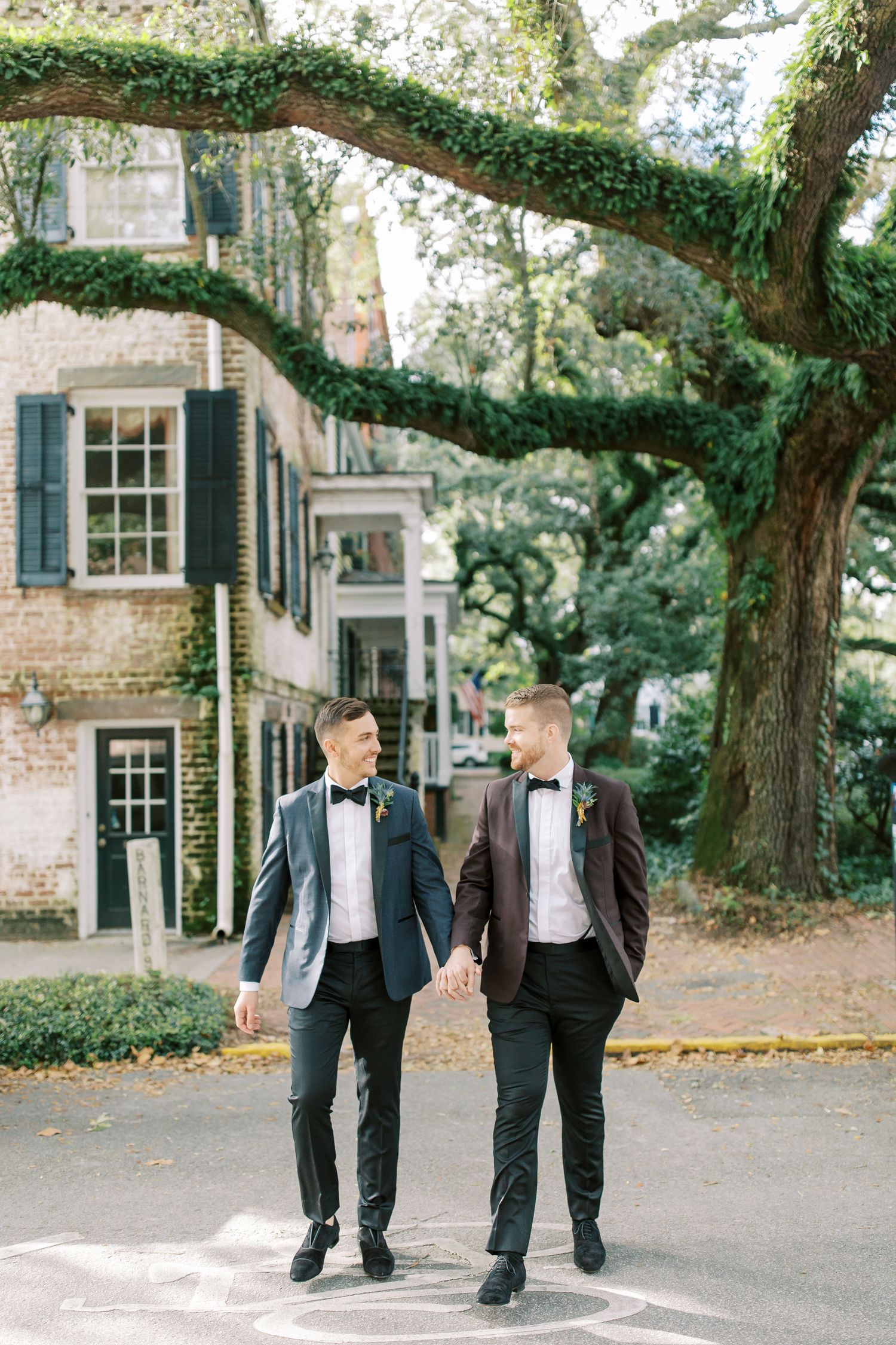 Two people in formal tuxedos walking together on a Savannah street with historic architecture and Spanish moss.