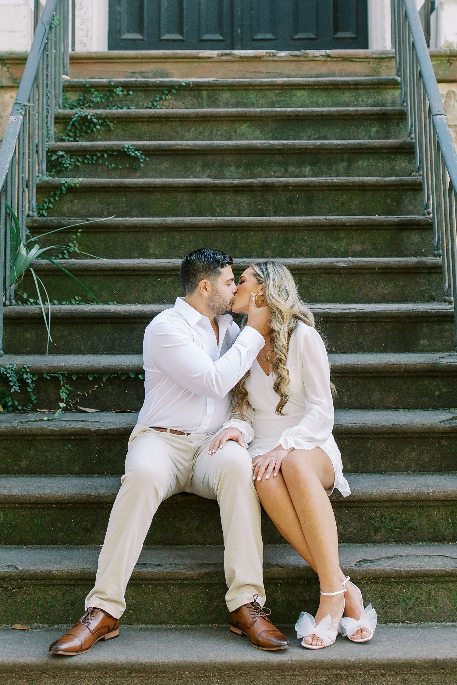 Romantic couple sitting on outdoor steps in matching white outfits share an intimate moment during engagement photoshoot.