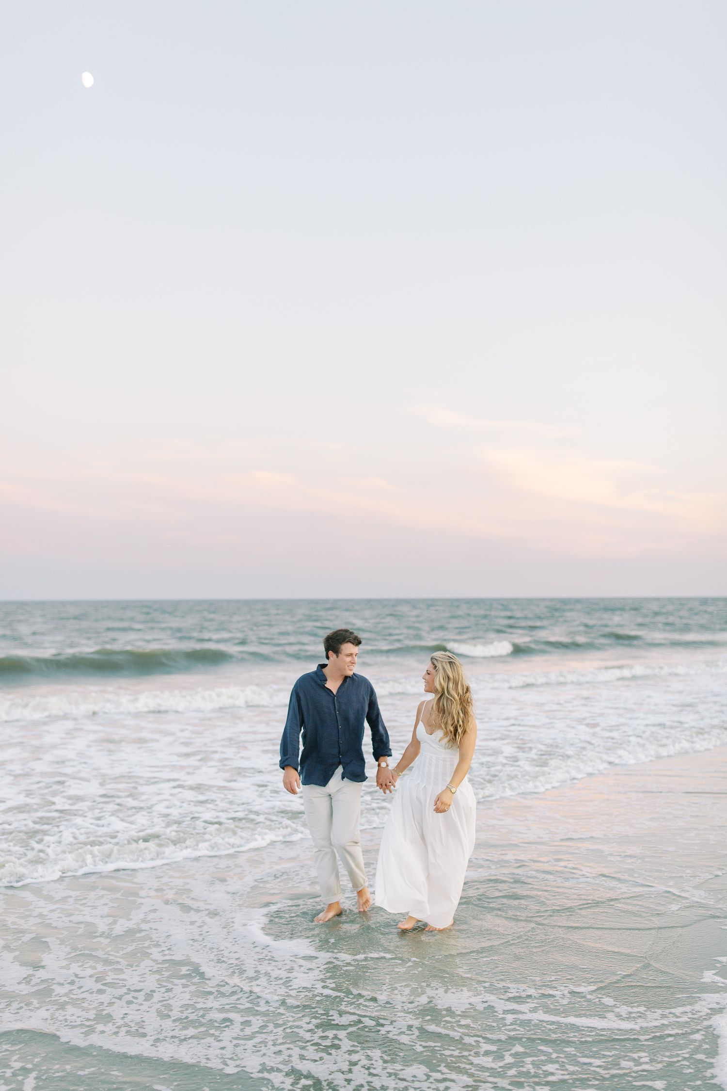 A couple walks hand in hand along the shoreline at sunset with waves lapping at their feet on St. Simons.
