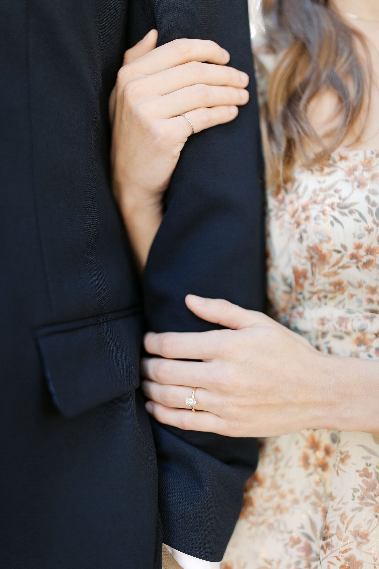 Close-up of hands showing an engagement ring against a navy suit jacket and floral dress.