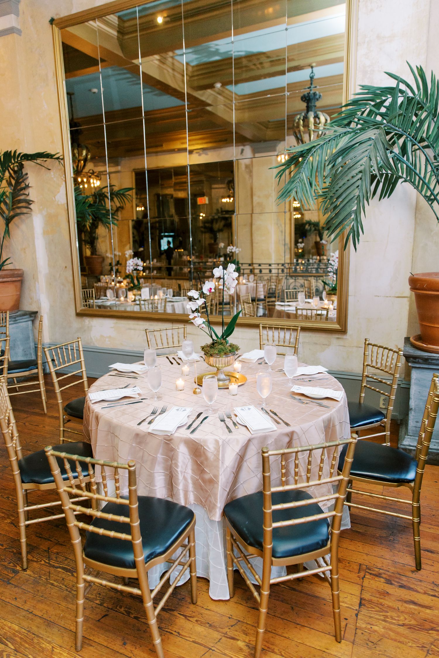 Elegant wedding reception table with gold Chiavari chairs and white tablecloth in a mirrored dining room with palm decor.