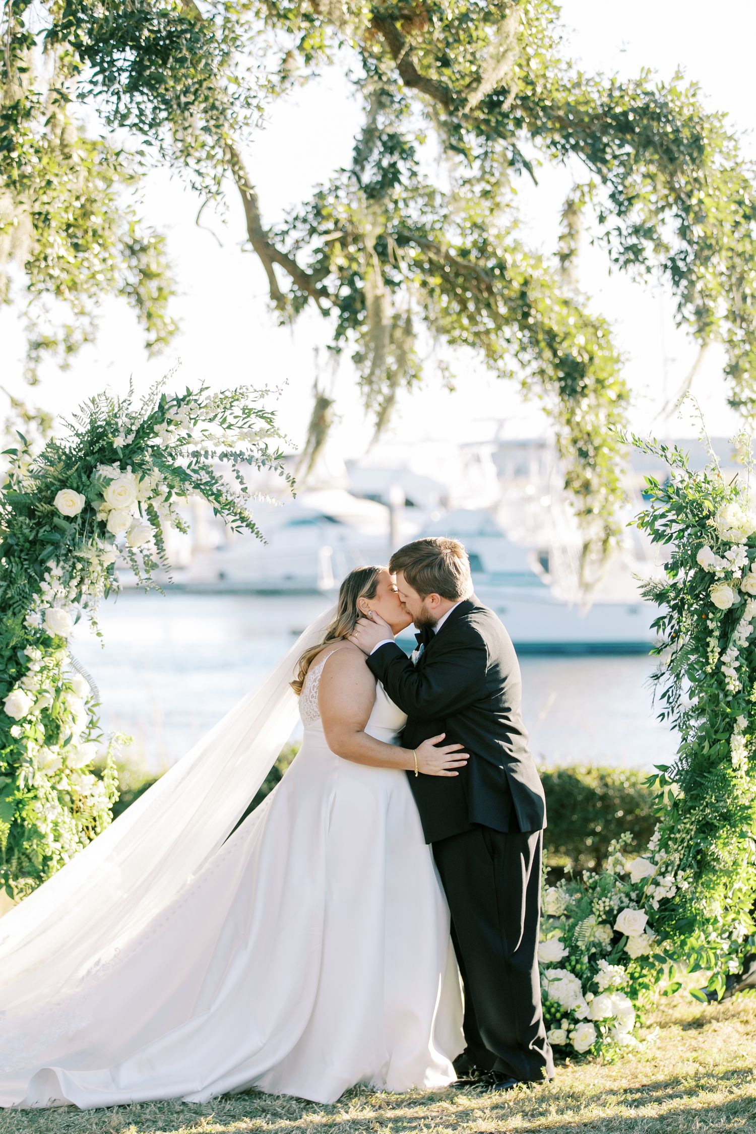 A romantic outdoor wedding ceremony under a floral arch with boats visible in the marina background.