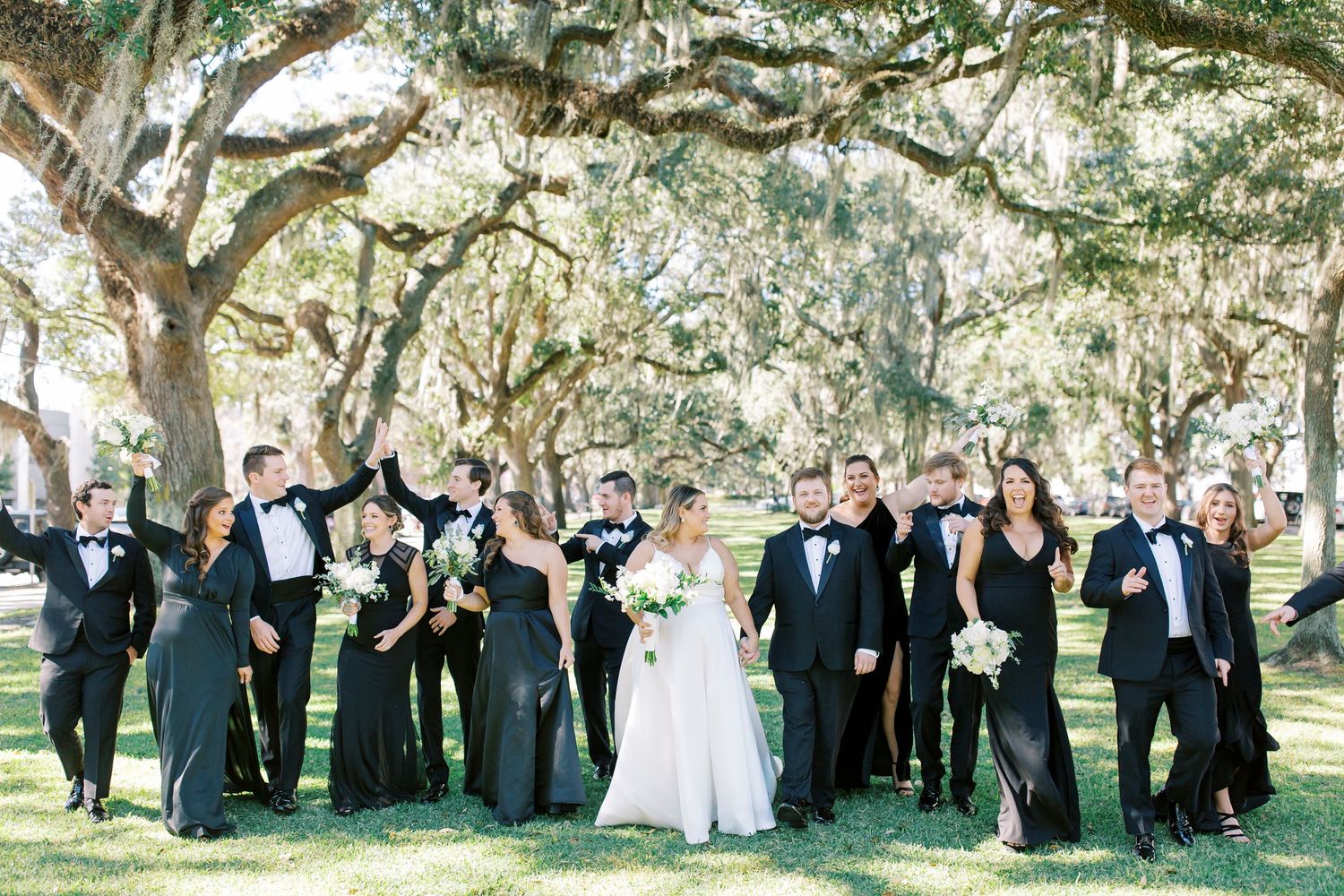 Wedding party in black formal attire poses under trees draped with Spanish moss.