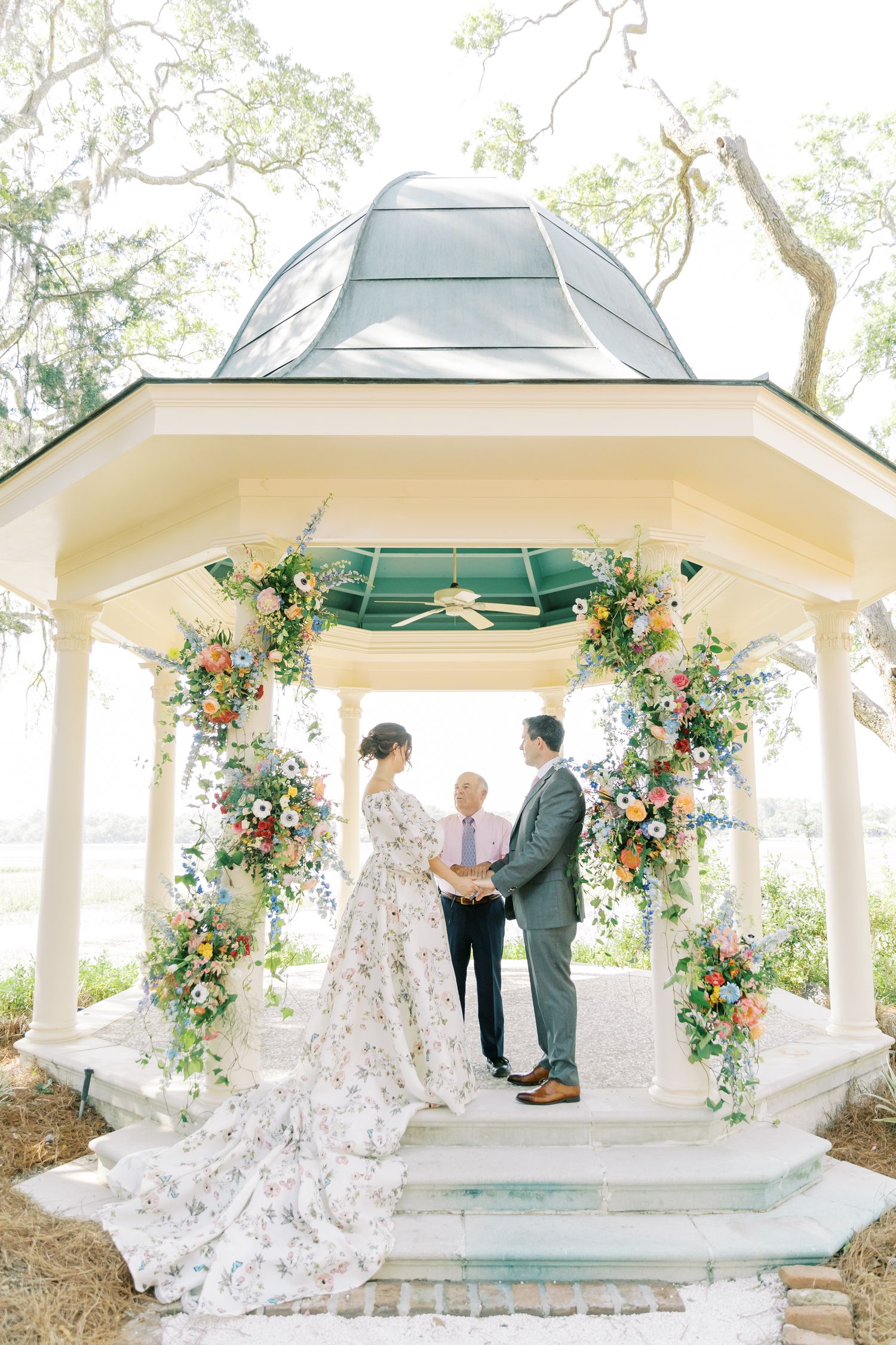 A wedding ceremony takes place in a white gazebo decorated with romantic floral arrangements and greenery.
