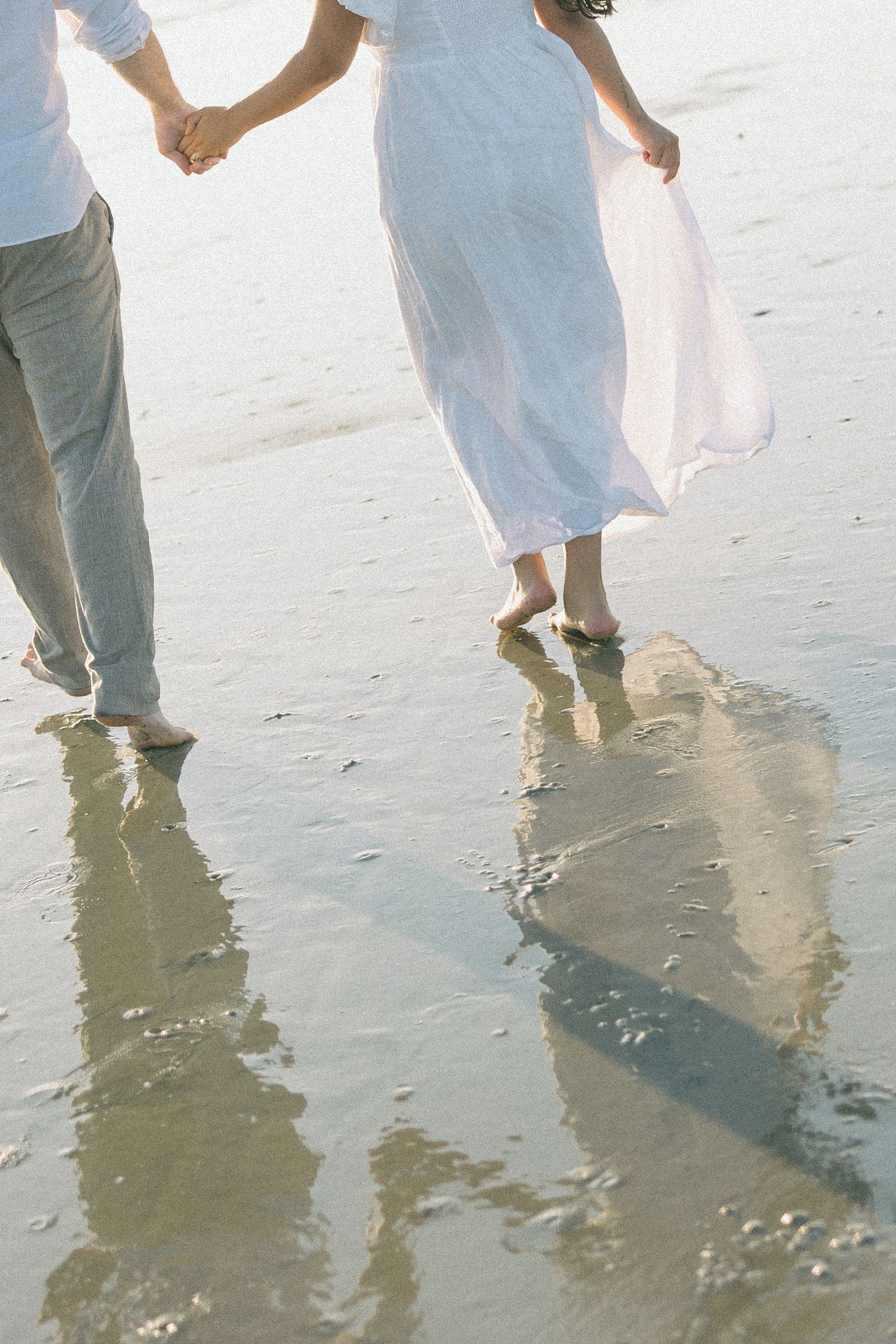 Feet of people standing in shallow ocean water at the beach with reflections on wet sand.
