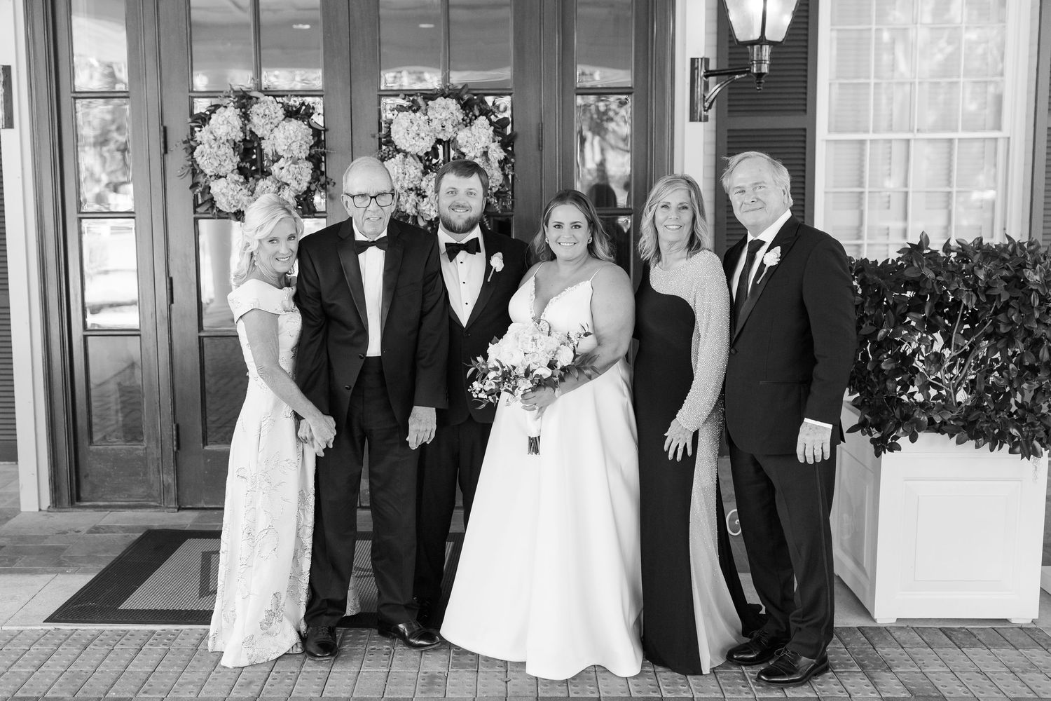 Wedding party in formal attire posing in front of decorated entrance with floral wreaths.