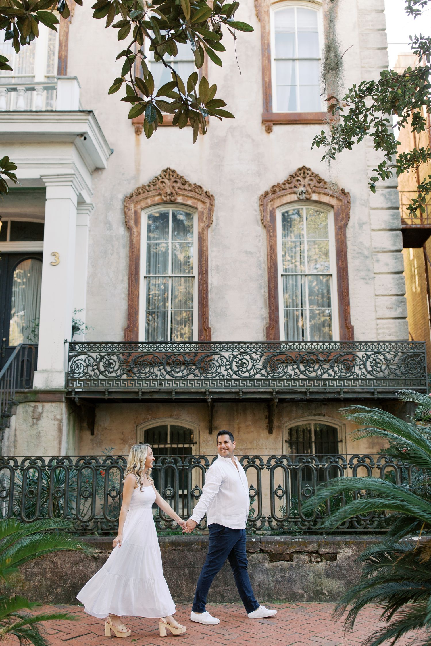 An elegant couple stands in front of an ornate Victorian-style building with magnolia branches and wrought iron railings in Savannah.