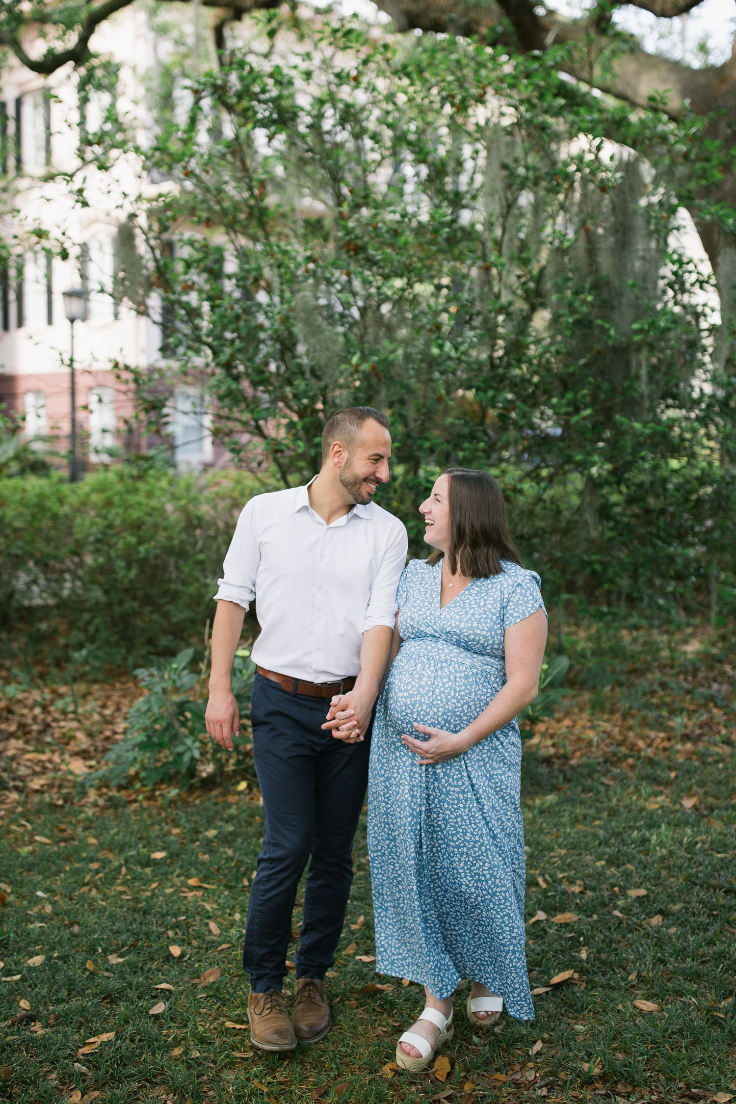 A couple embraces outdoors in a garden setting wearing casual attire and smiling at each other.