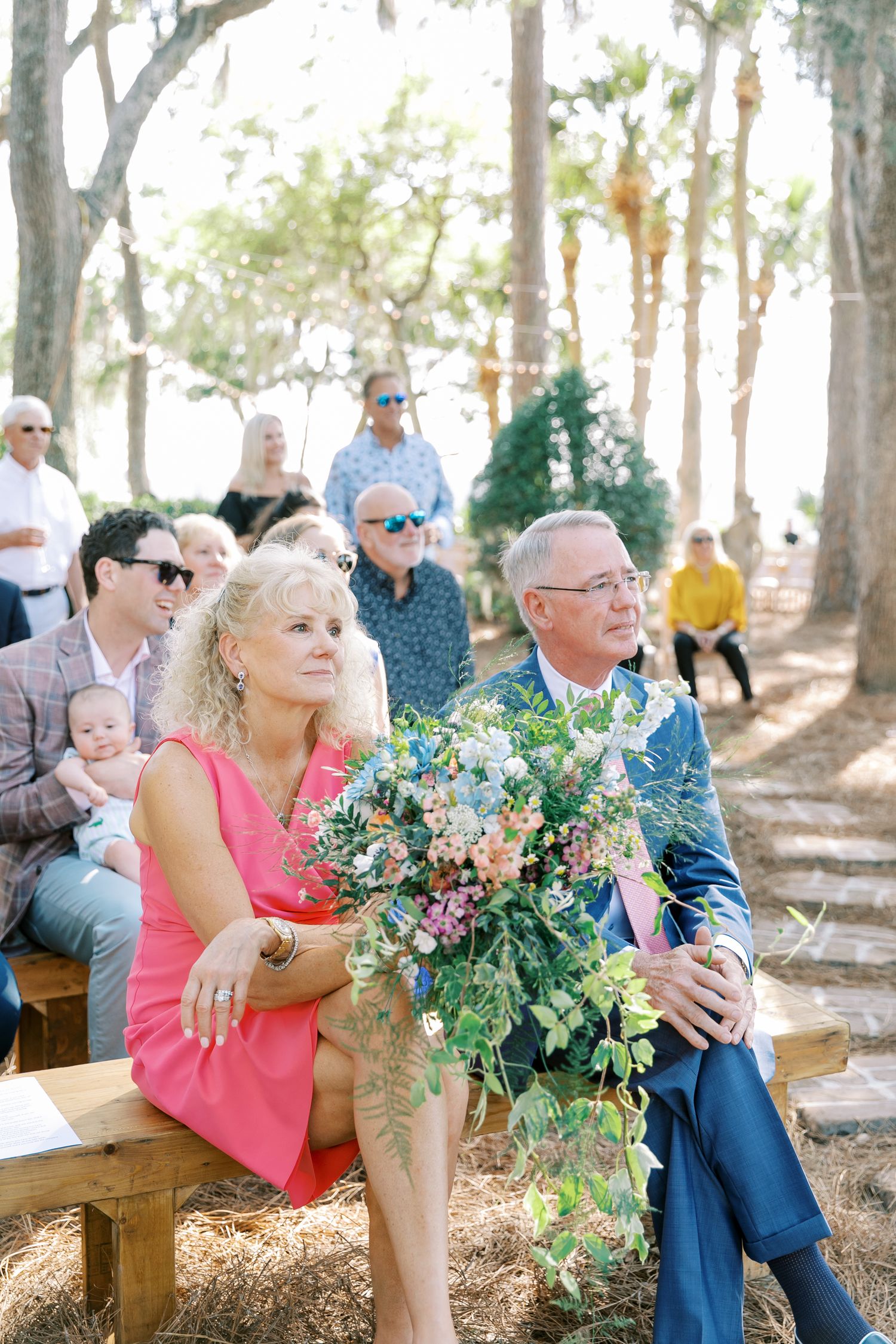 Guests seated on wooden benches at an outdoor wedding ceremony beneath towering trees.