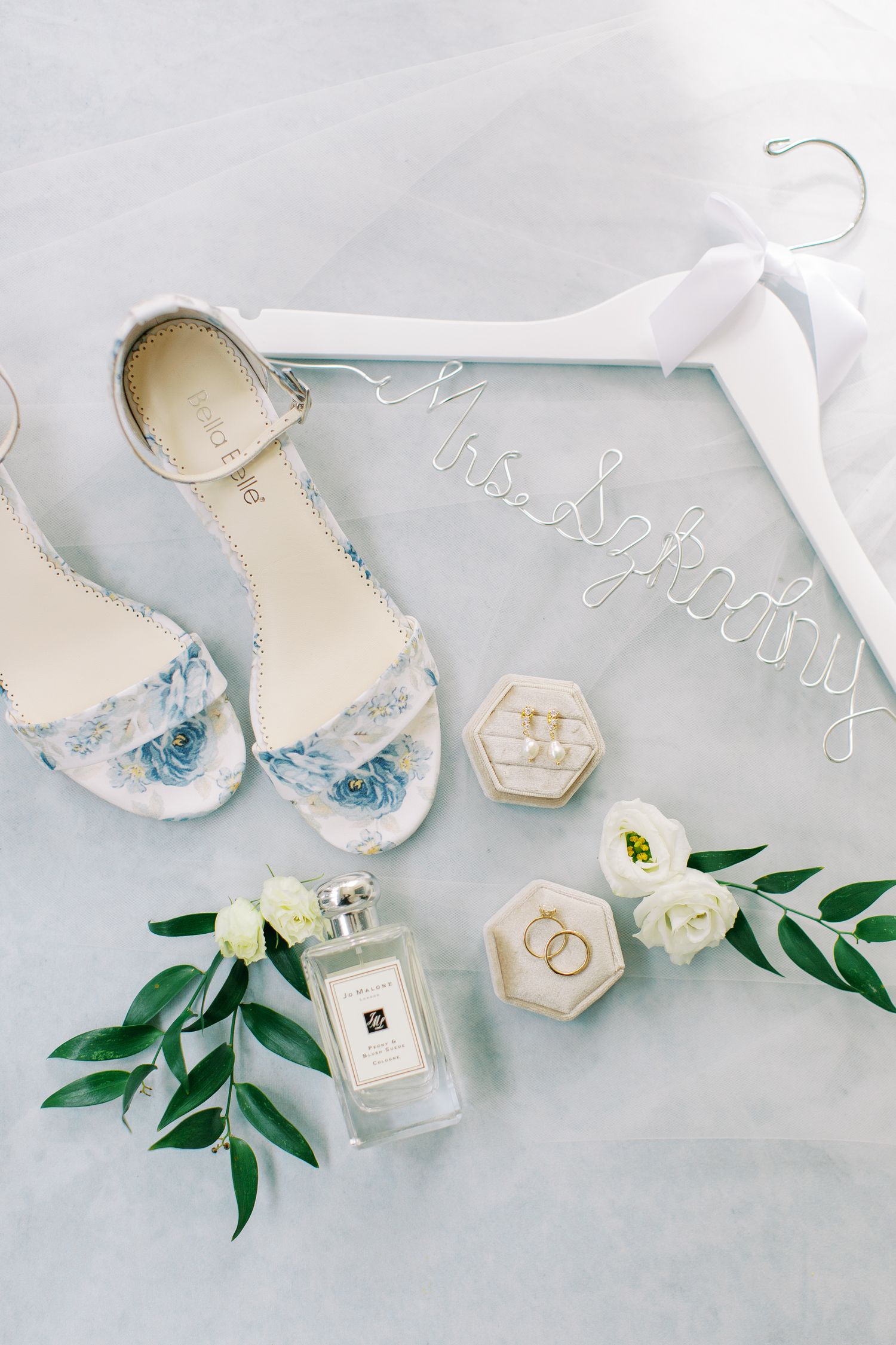 Wedding accessories including blue shoes, perfume bottle, and personalized hanger on light background.