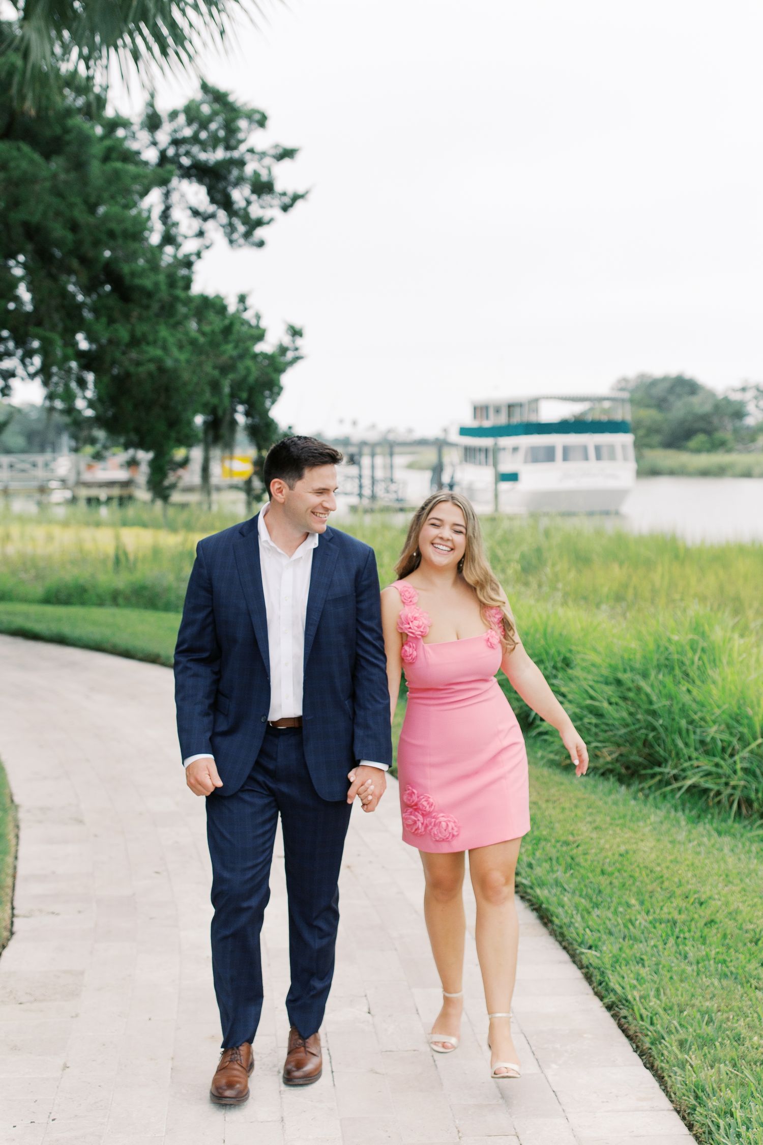 A couple walking on a path near water, wearing formal attire with a navy suit and pink dress on a sunny day.