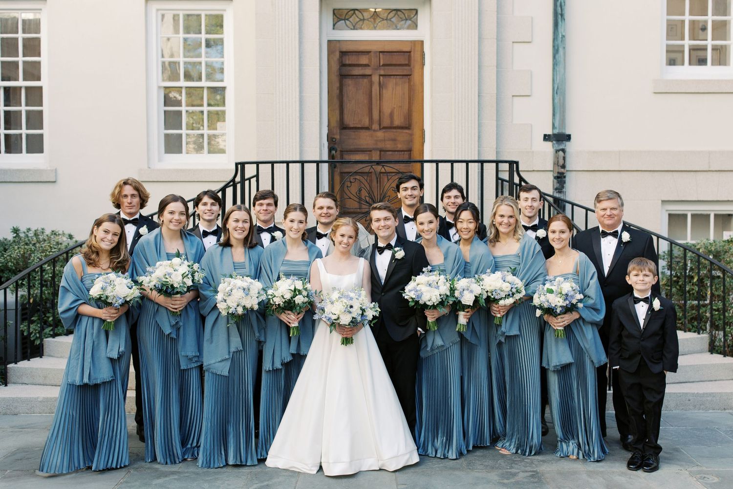 Wedding party in matching dusty blue bridesmaid dresses and formal attire pose on stone steps of traditional building.