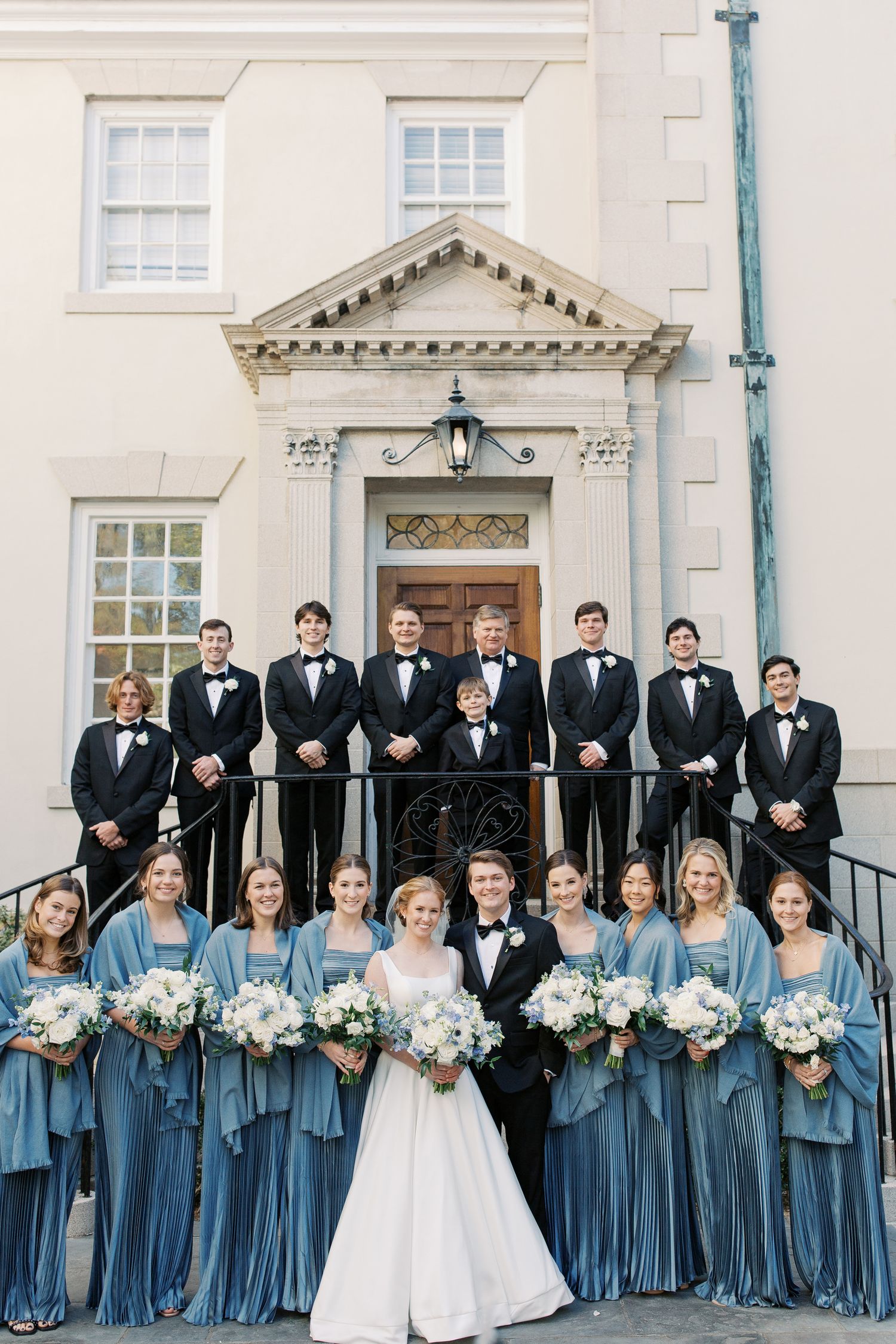 Wedding party poses on historic building steps with bridesmaids in blue and groomsmen in black tuxedos.