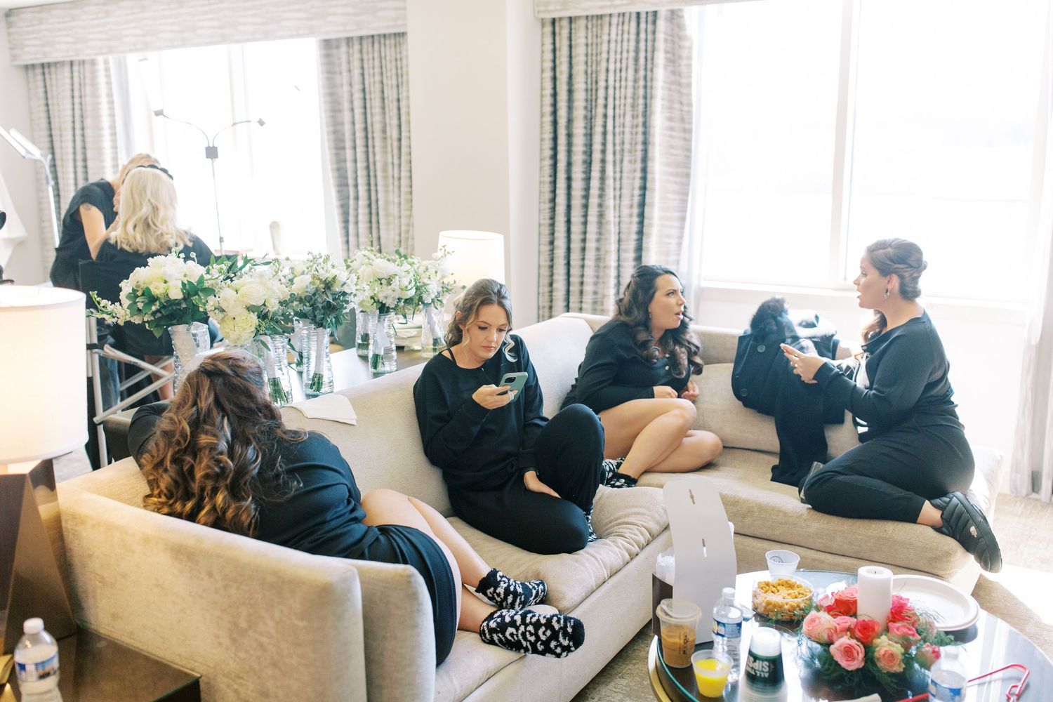 People gather in casual attire sitting on beige furniture during an indoor social gathering with drinks and decor visible.