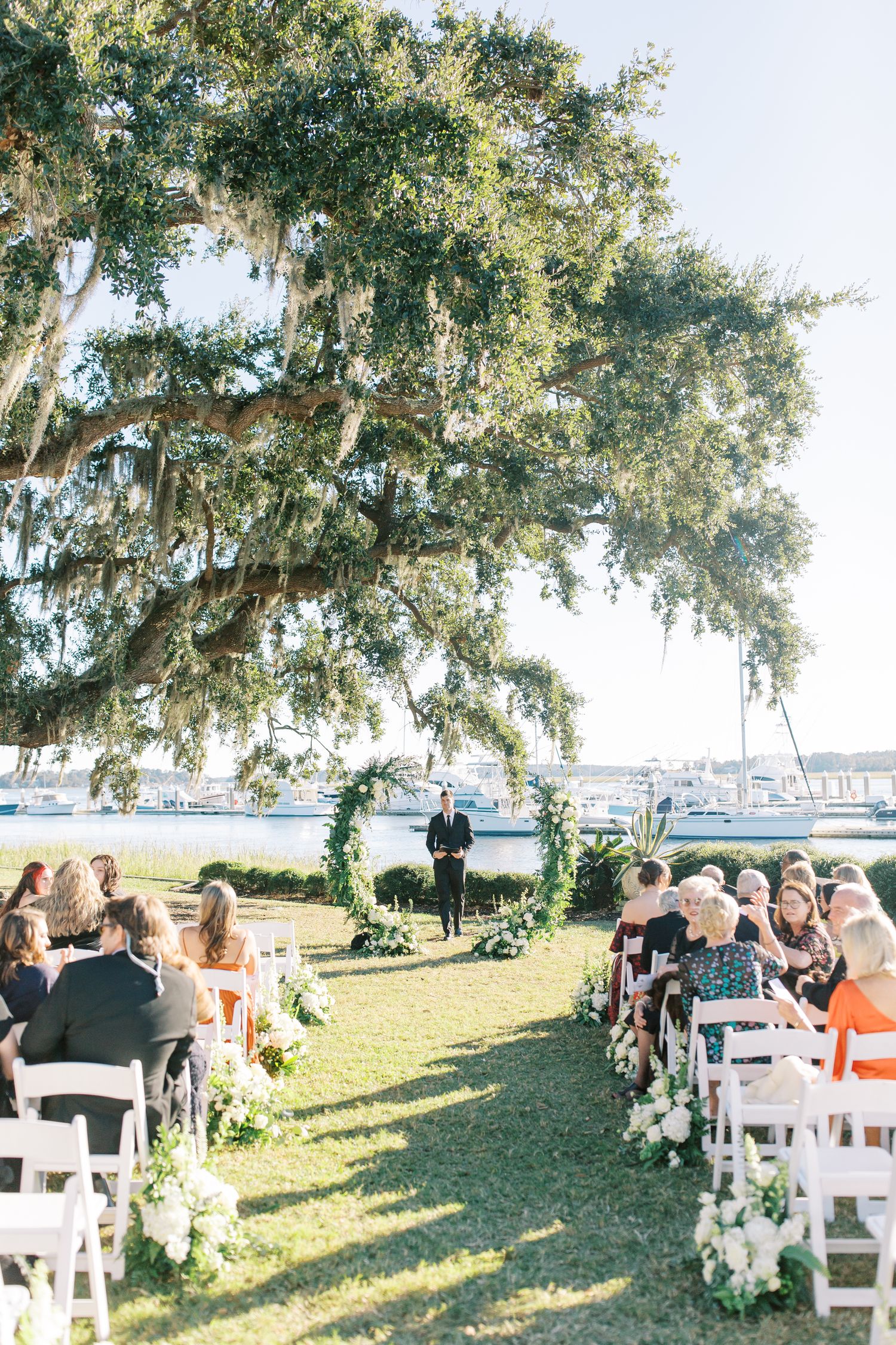 Outdoor wedding ceremony setup under a majestic oak tree with Spanish moss and white chairs.