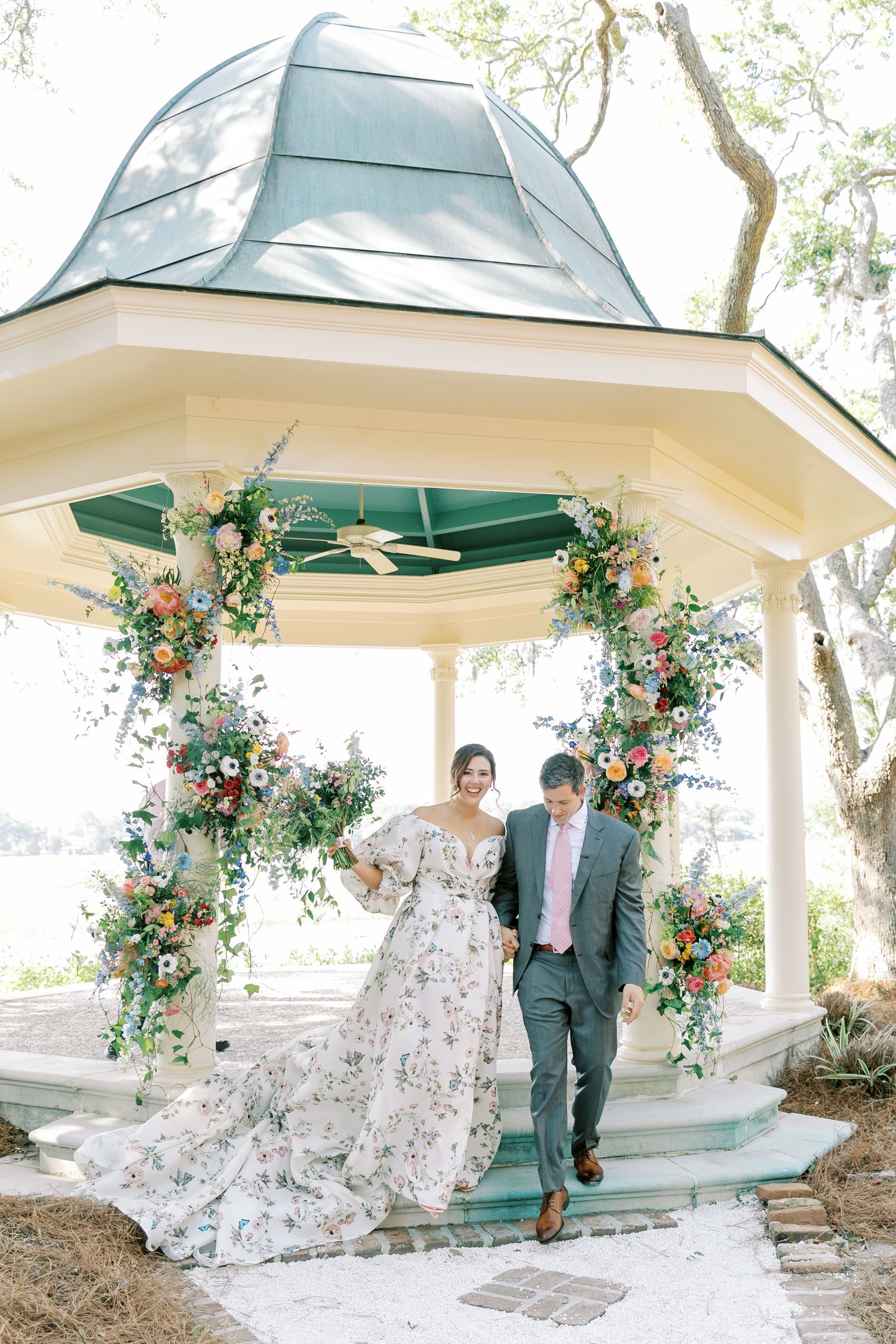 A couple shares a romantic moment under a white gazebo decorated with colorful floral arrangements and greenery.