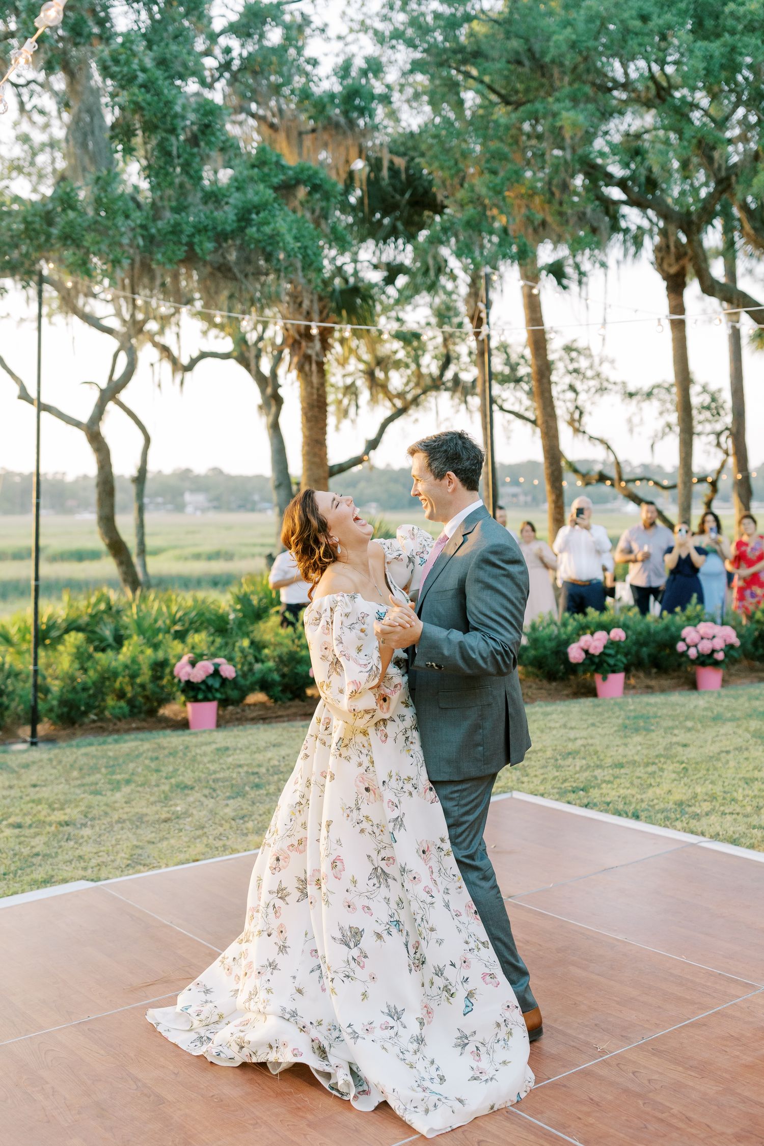 Wedding couple dances on outdoor terrace surrounded by trees and pink floral decorations.