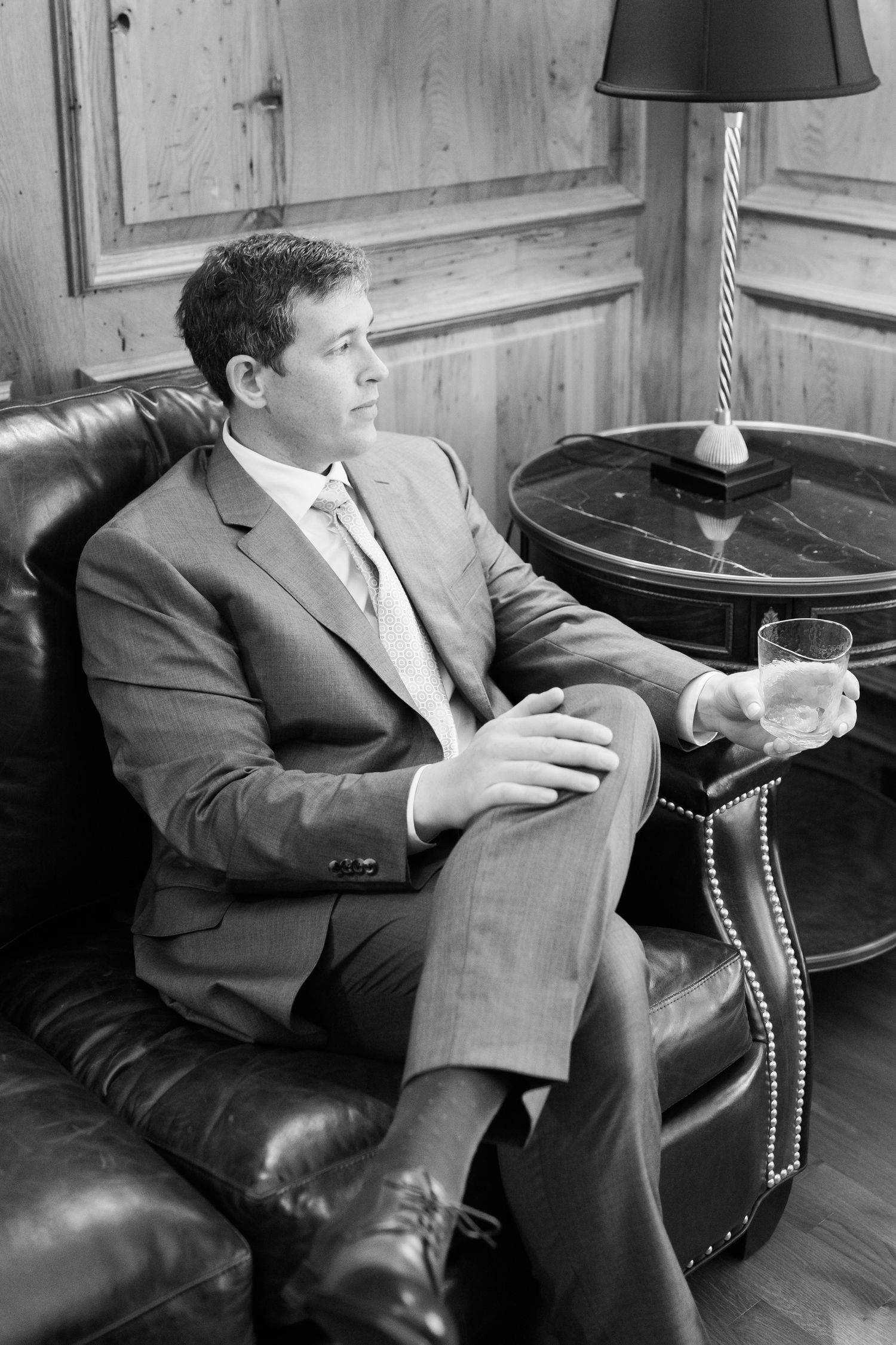 Vintage black and white photo of a man in suit relaxing in leather chair with drink next to side table and lamp.