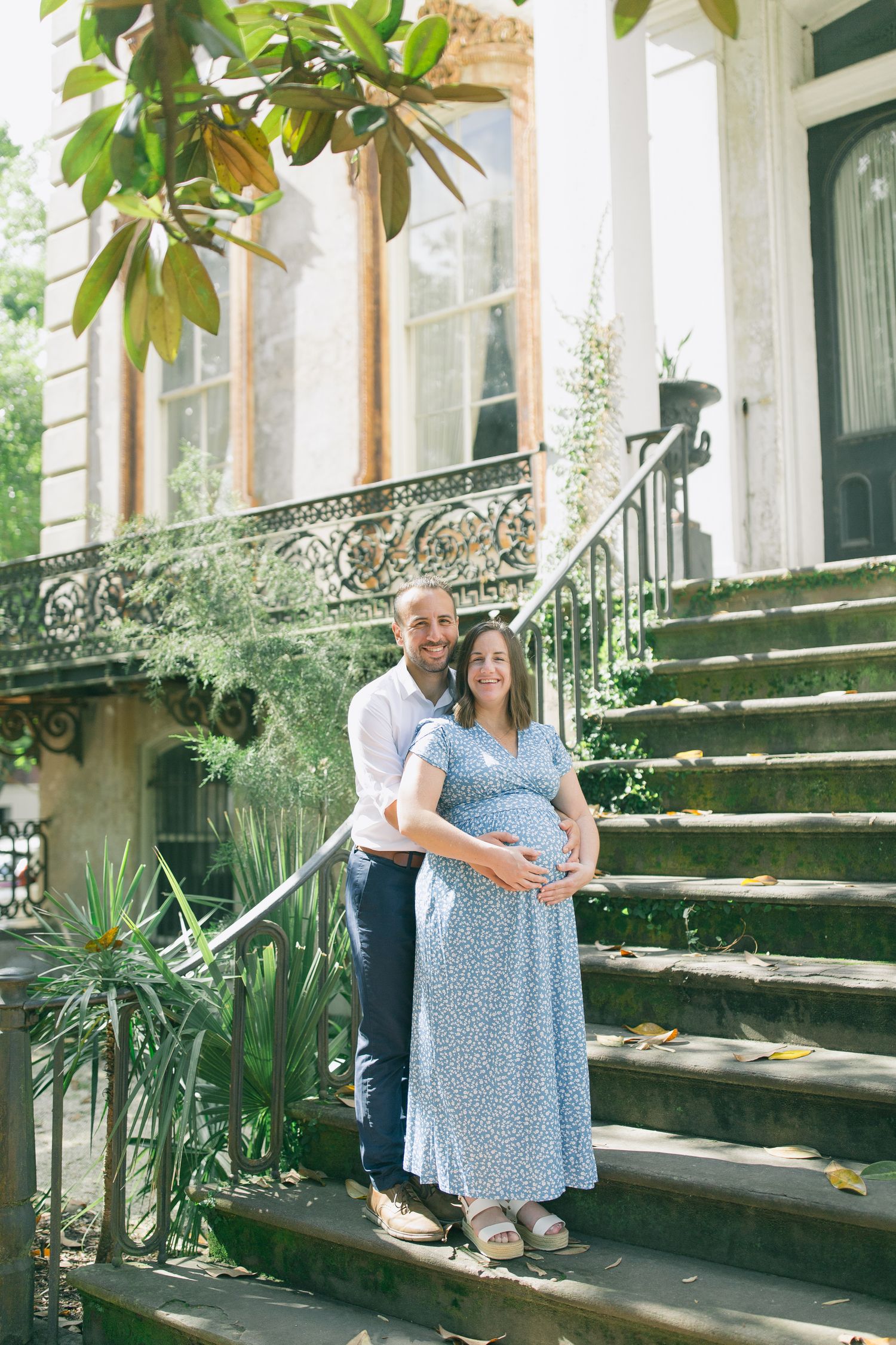A couple poses affectionately on historic stone steps surrounded by greenery and magnolia trees.