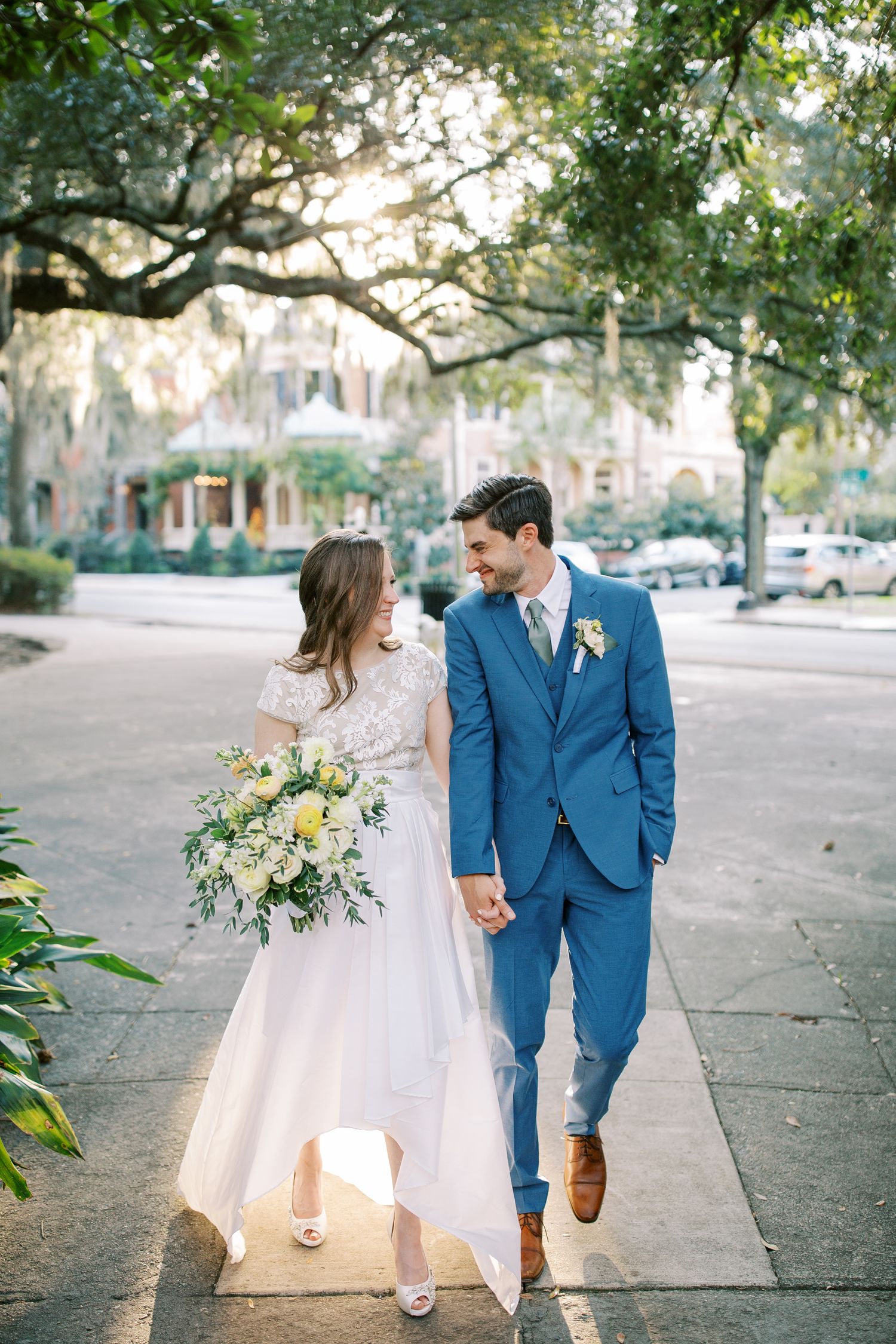 Newlyweds walk hand in hand down a tree-lined sidewalk with the bride carrying a white floral bouquet.