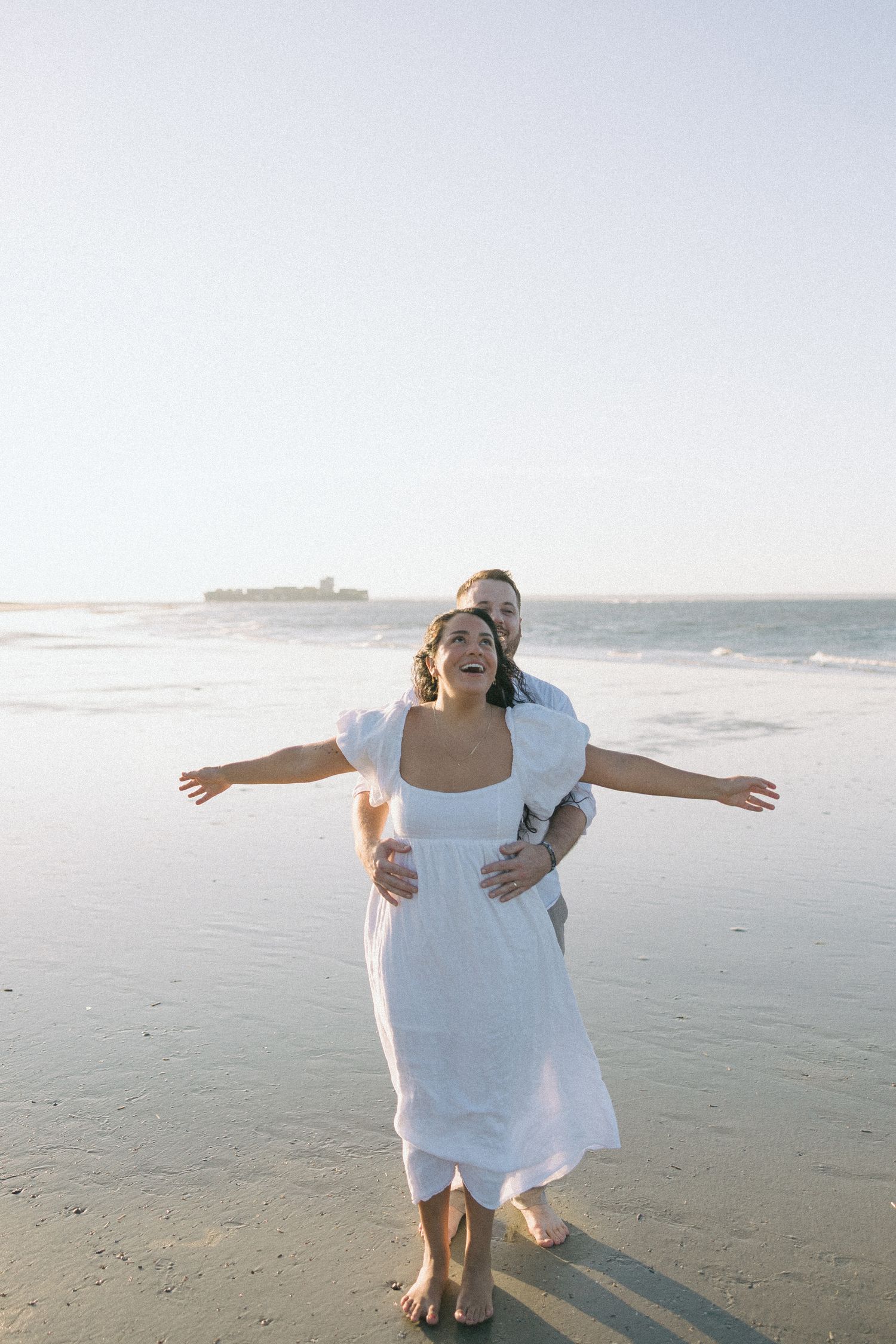 A couple embraces on a sunny beach with waves and ocean in the background as sunlight casts long shadows on the sand.