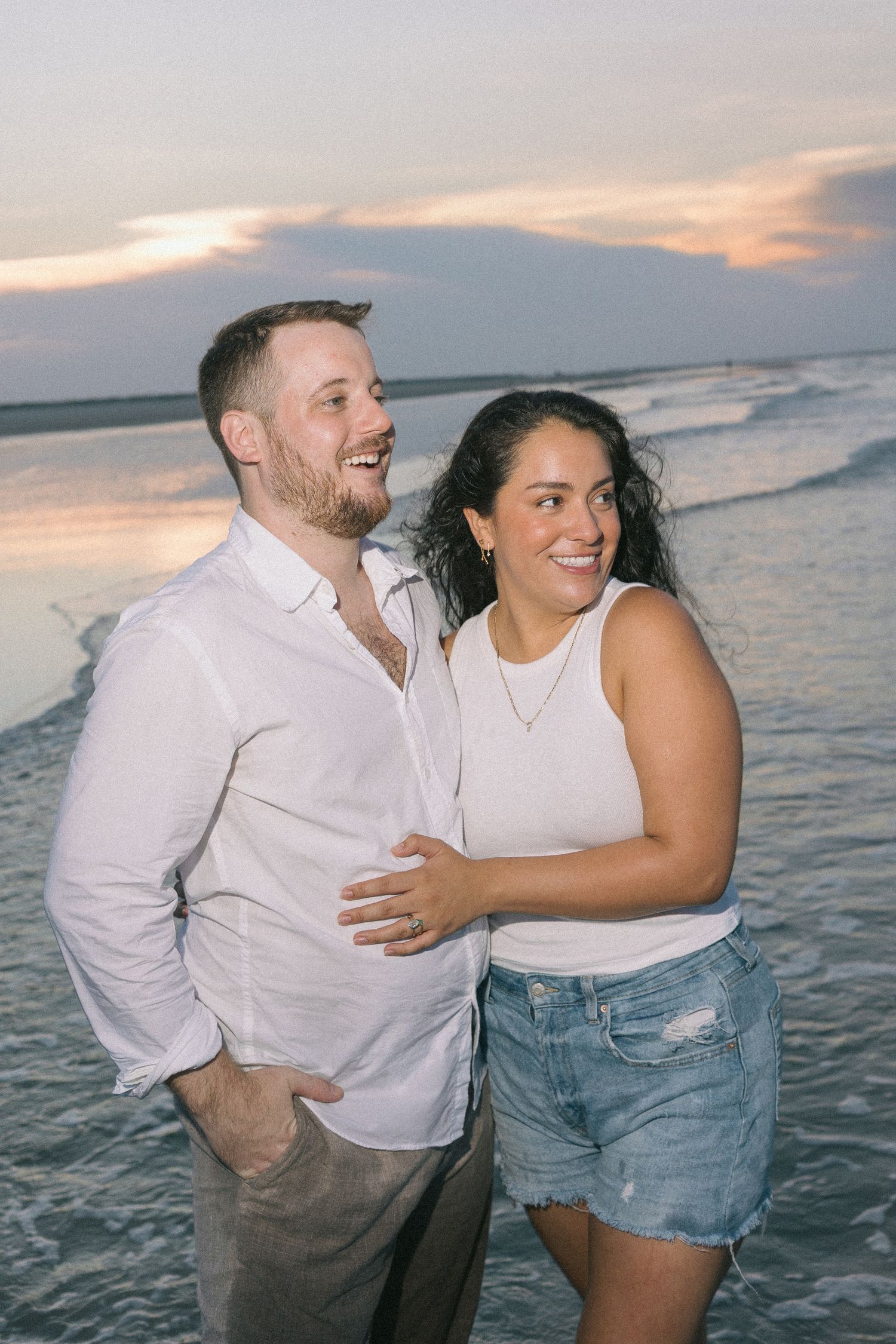 Couple laughing and embracing on beach at sunset with ocean waves in background.