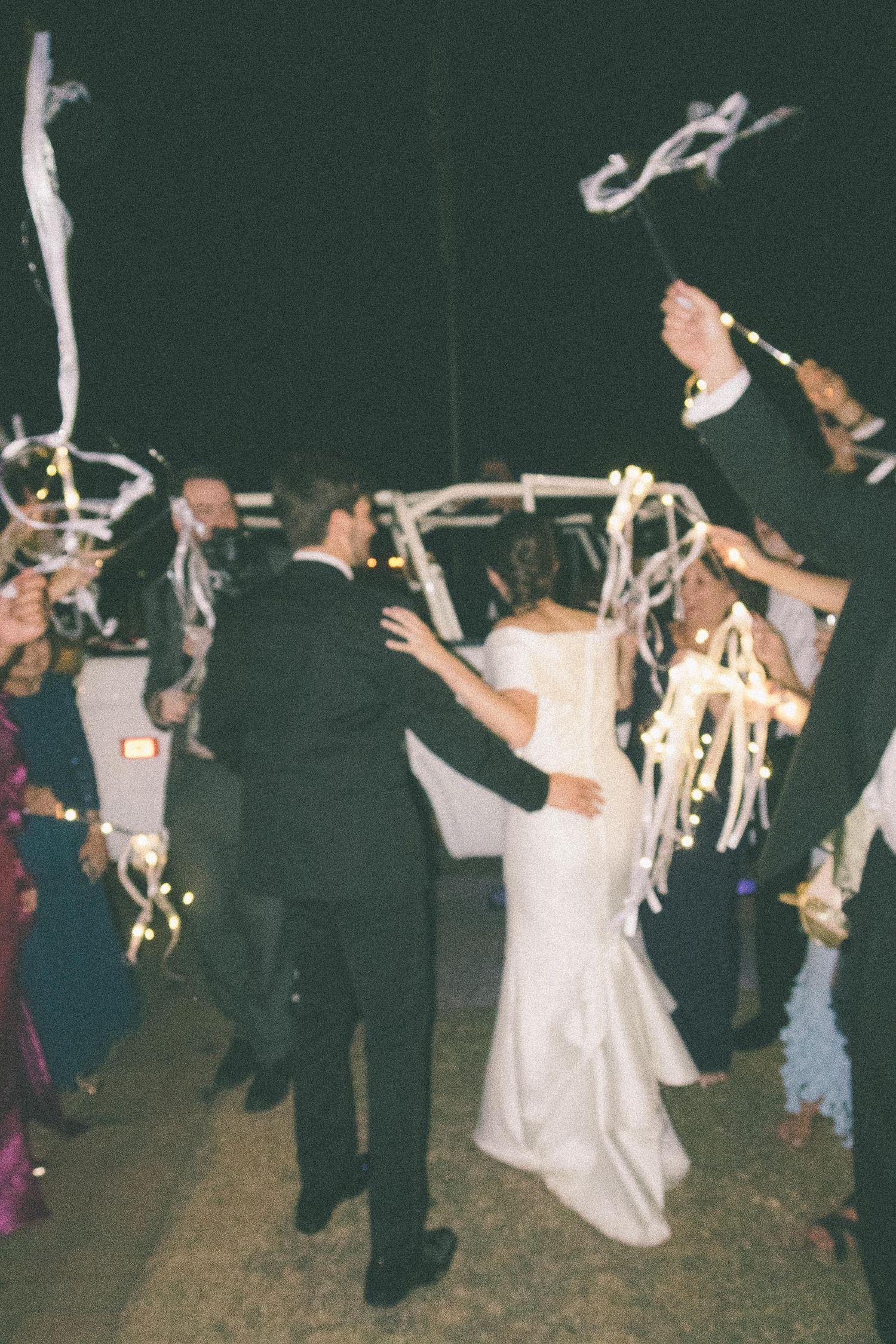 Wedding guests dance and celebrate at a nighttime reception with people in formal attire and white dresses visible.