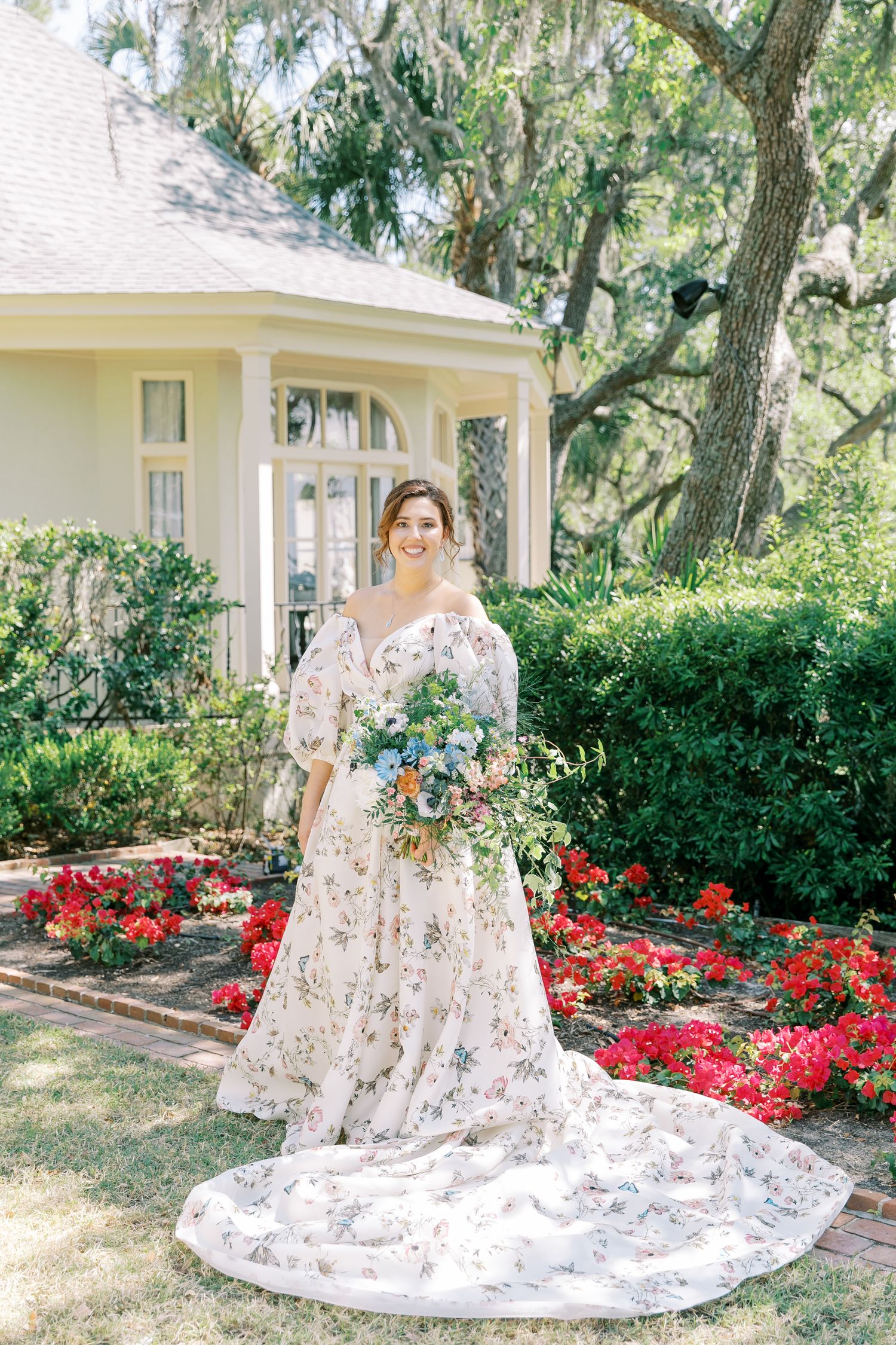 A floral wedding dress with long train photographed in front of a cream colored house with red flowers in the garden.
