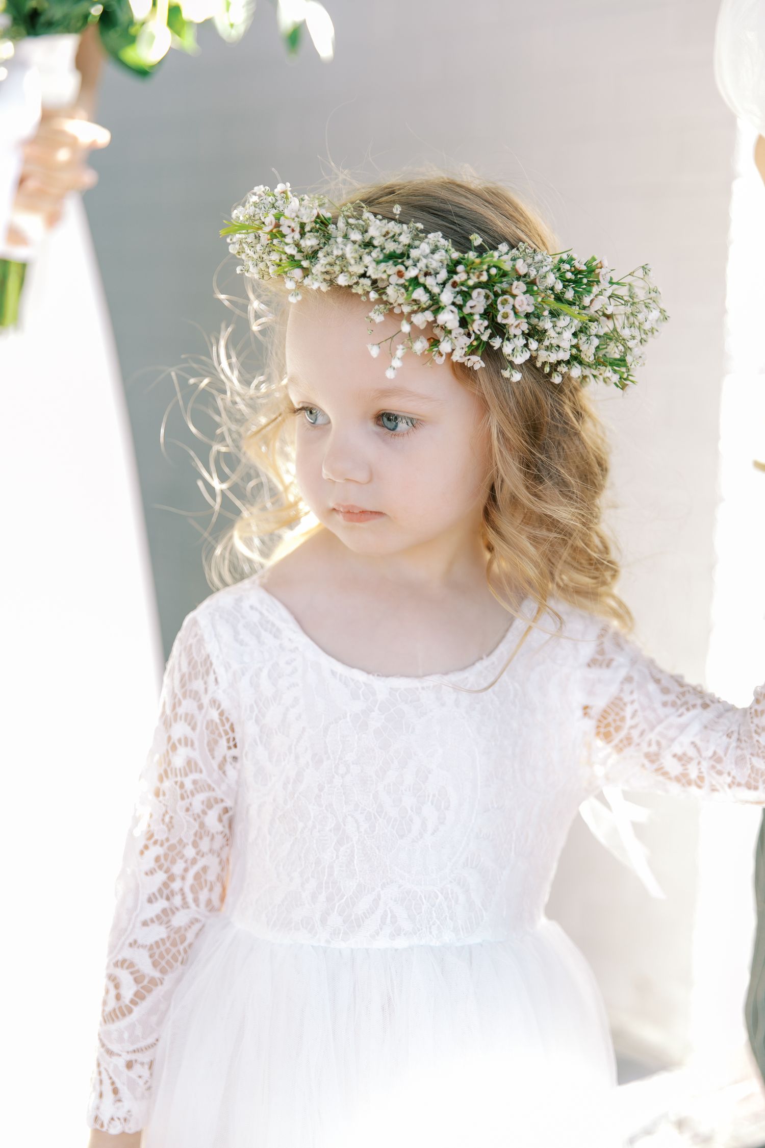 A young flower girl in white lace dress wears a delicate baby's breath flower crown.