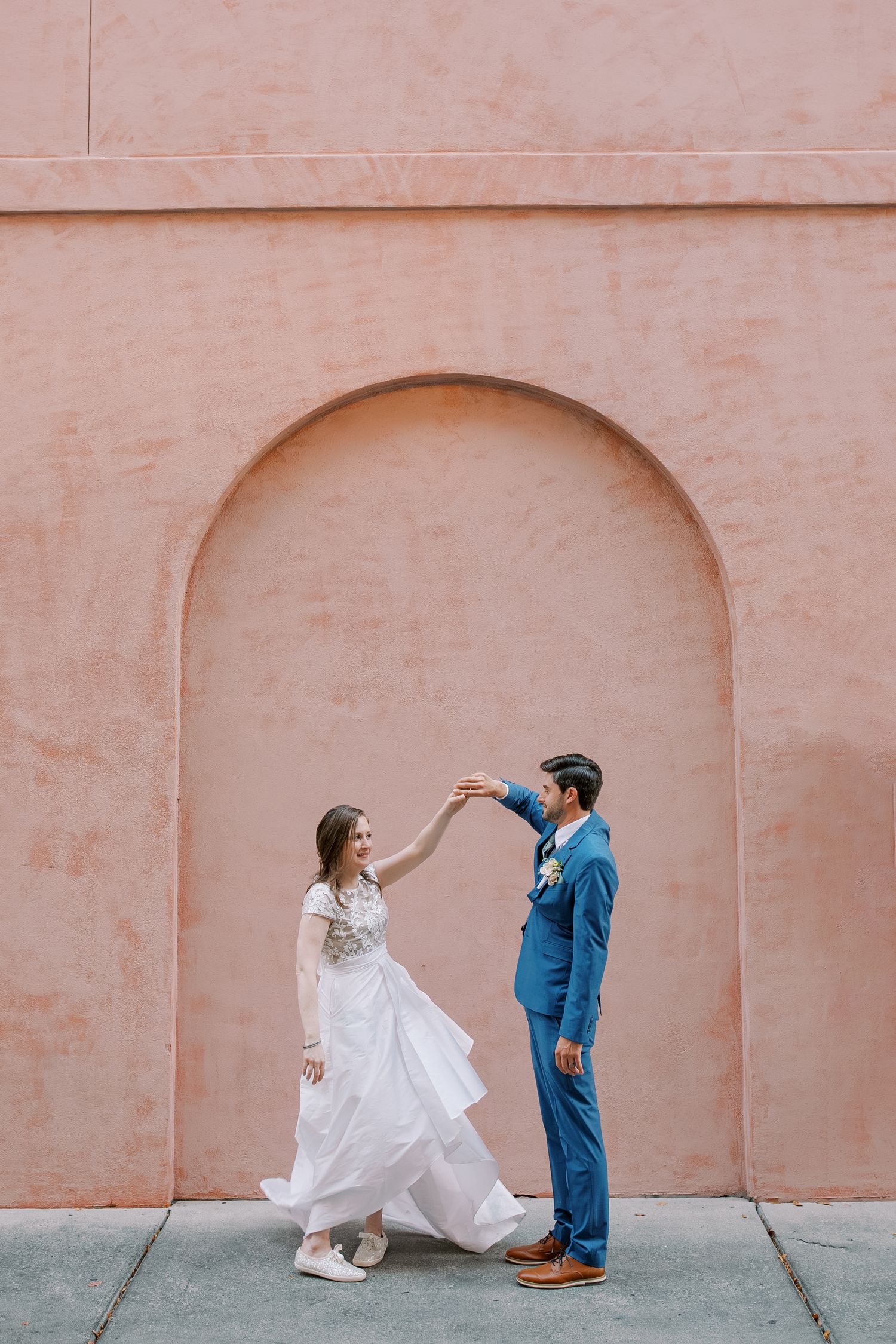 A playful wedding portrait under a pink arched doorway in an urban setting.