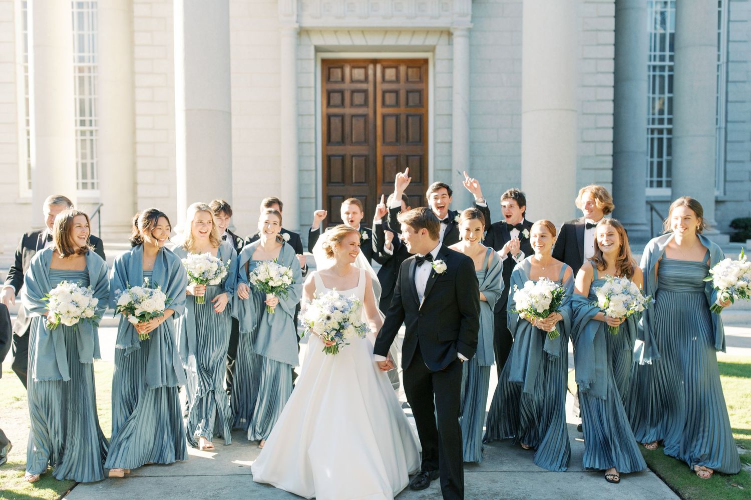 Wedding party in dusty blue bridesmaid dresses celebrating outside a grand white building.
