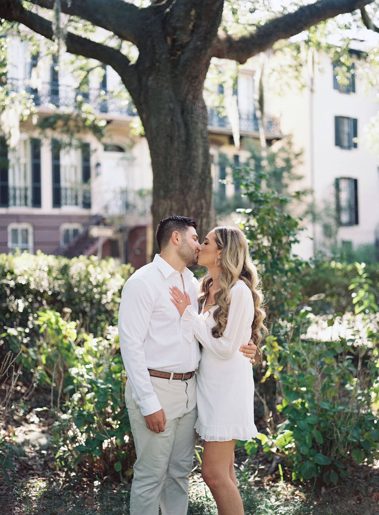 Romantic moment captured under large oak tree with historic building in background.