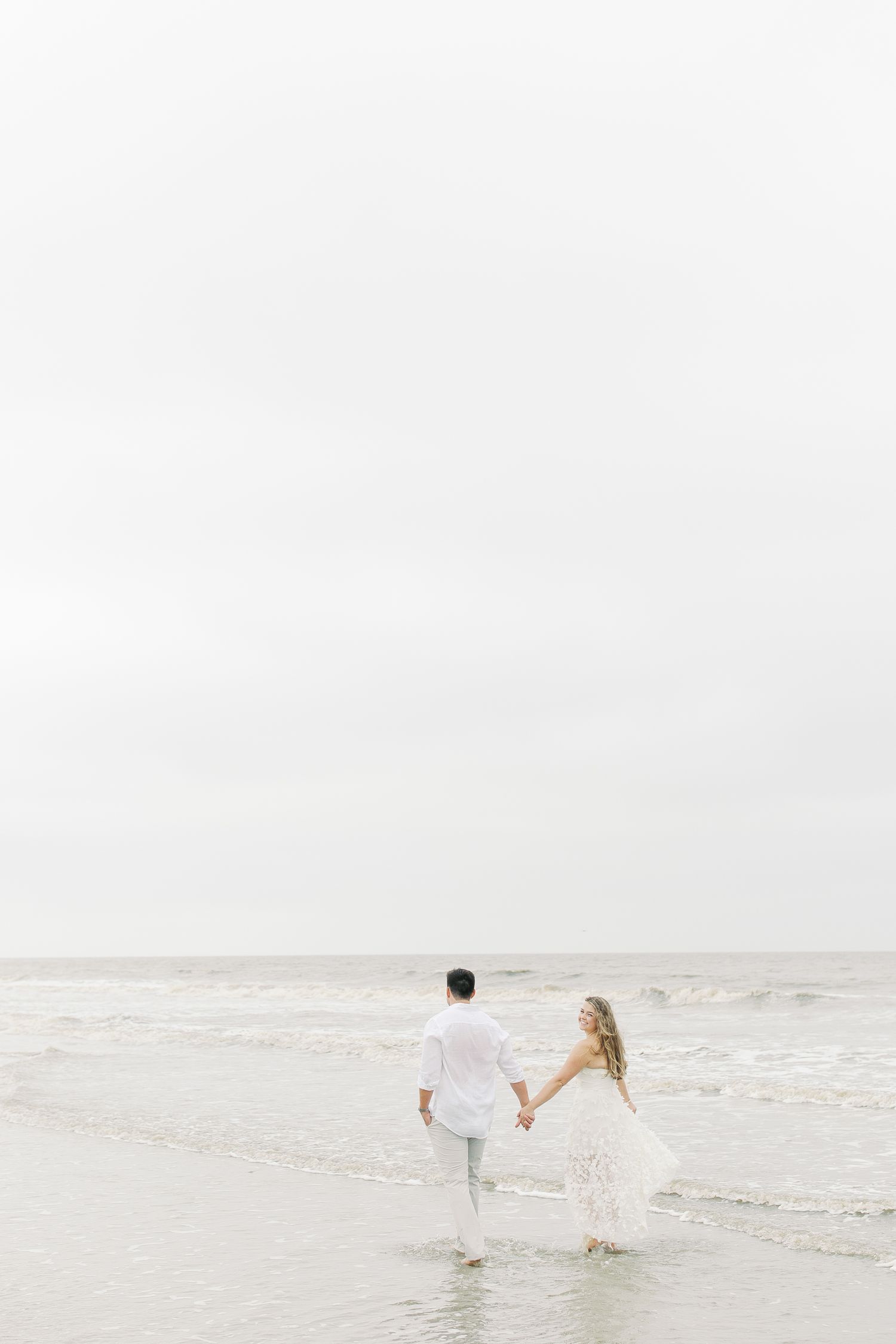 Couple in white attire walking along misty beach shore.